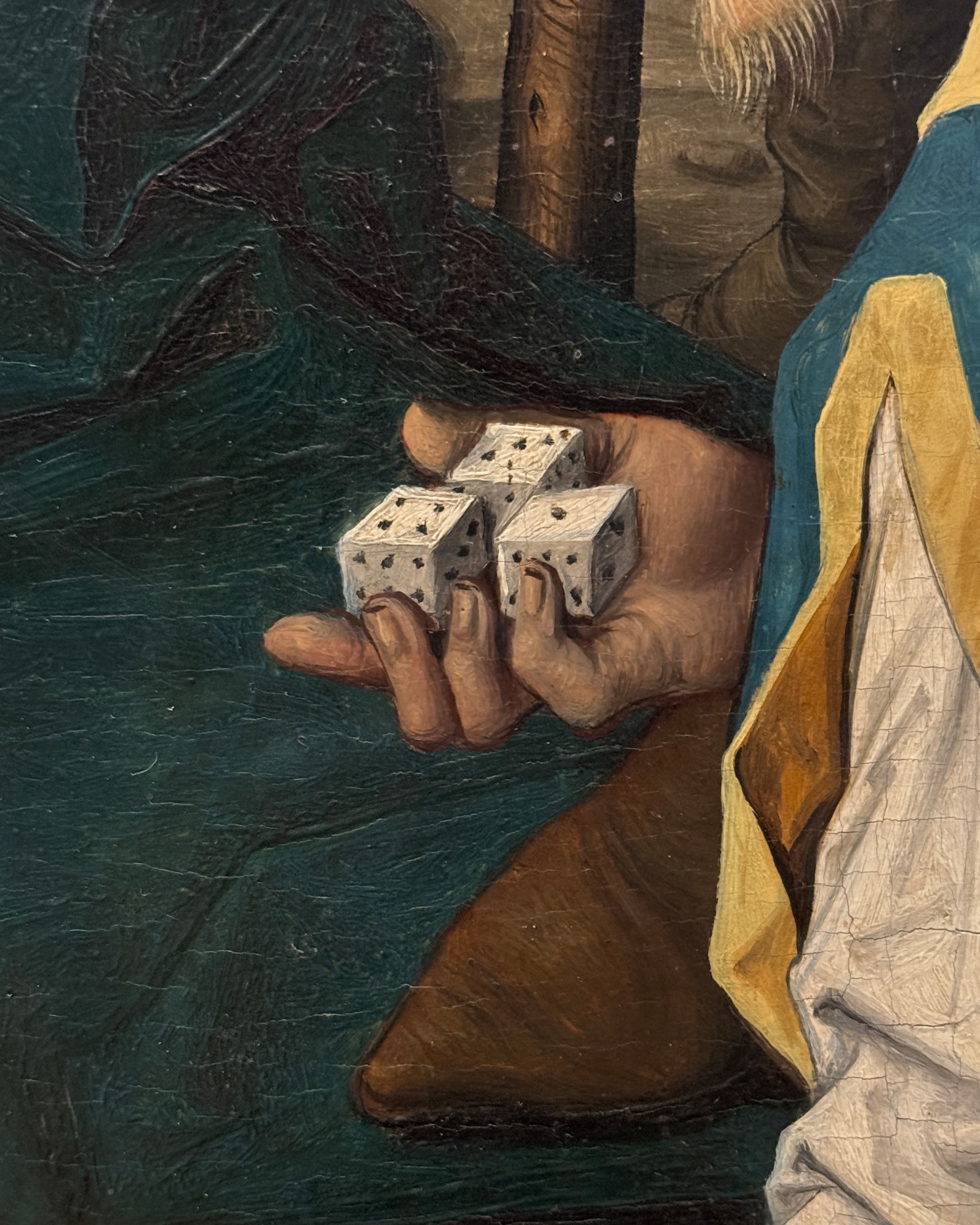 Eine Hand in einem blauen und gelben Ärmel hält drei weiße Würfel mit schwarzen Punkten. Das Bild ist ein Detail aus einem Gemälde mit sichtbaren Rissen in der Oberfläche. (KI-Alternativtext)