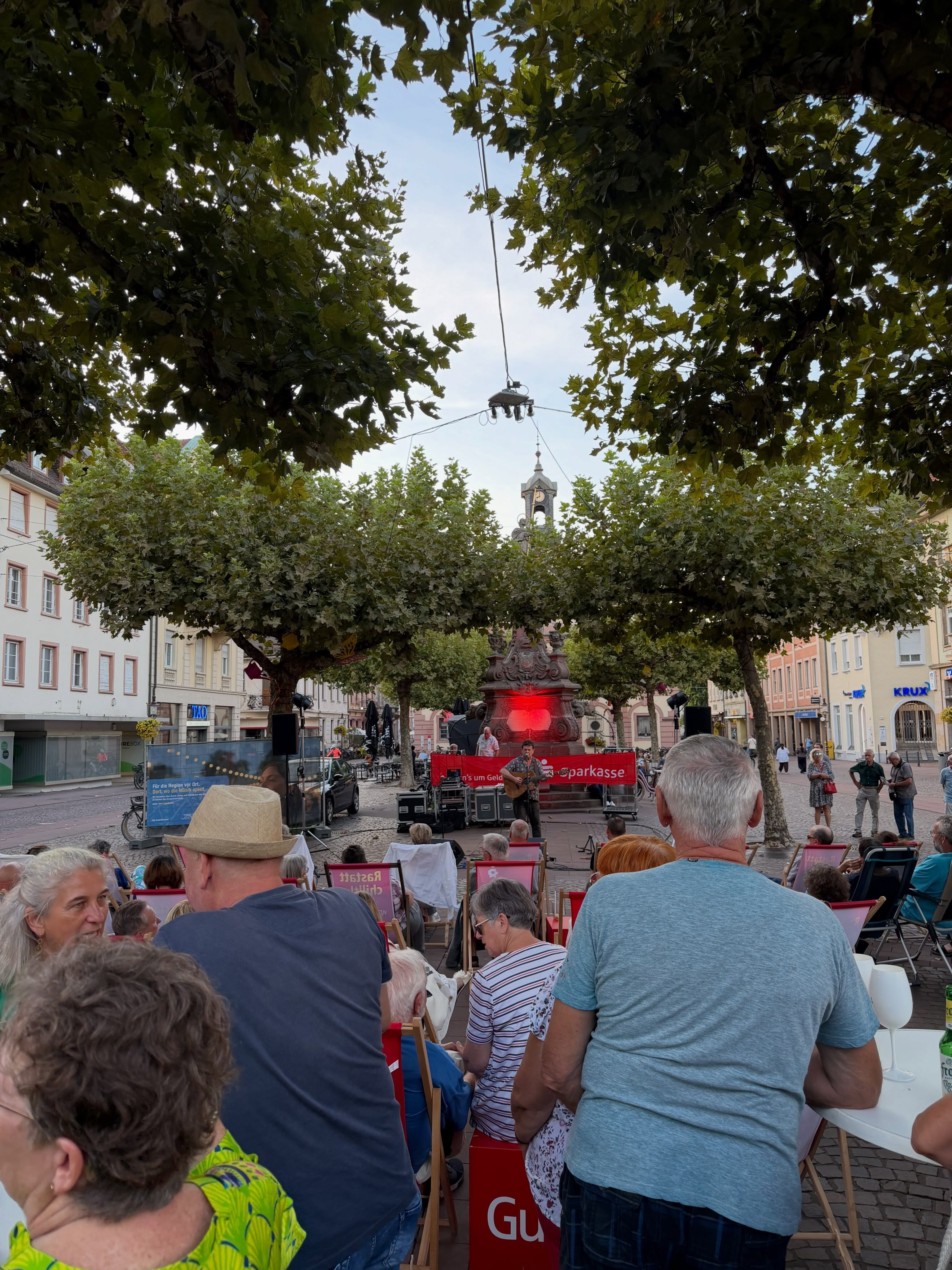 Ein Musiker spielt Gitarre auf einer Bühne auf einem Platz, während ein Publikum auf Klappstühlen zuschaut. Hinter der Bühne hängt ein rotes Banner mit den Schriftzügen "n's um Geld" und "parkasse". Im Hintergrund erhebt sich ein Brunnen mit roter Beleuchtung zwischen Bäumen und Gebäuden. (KI-Alternativtext)
