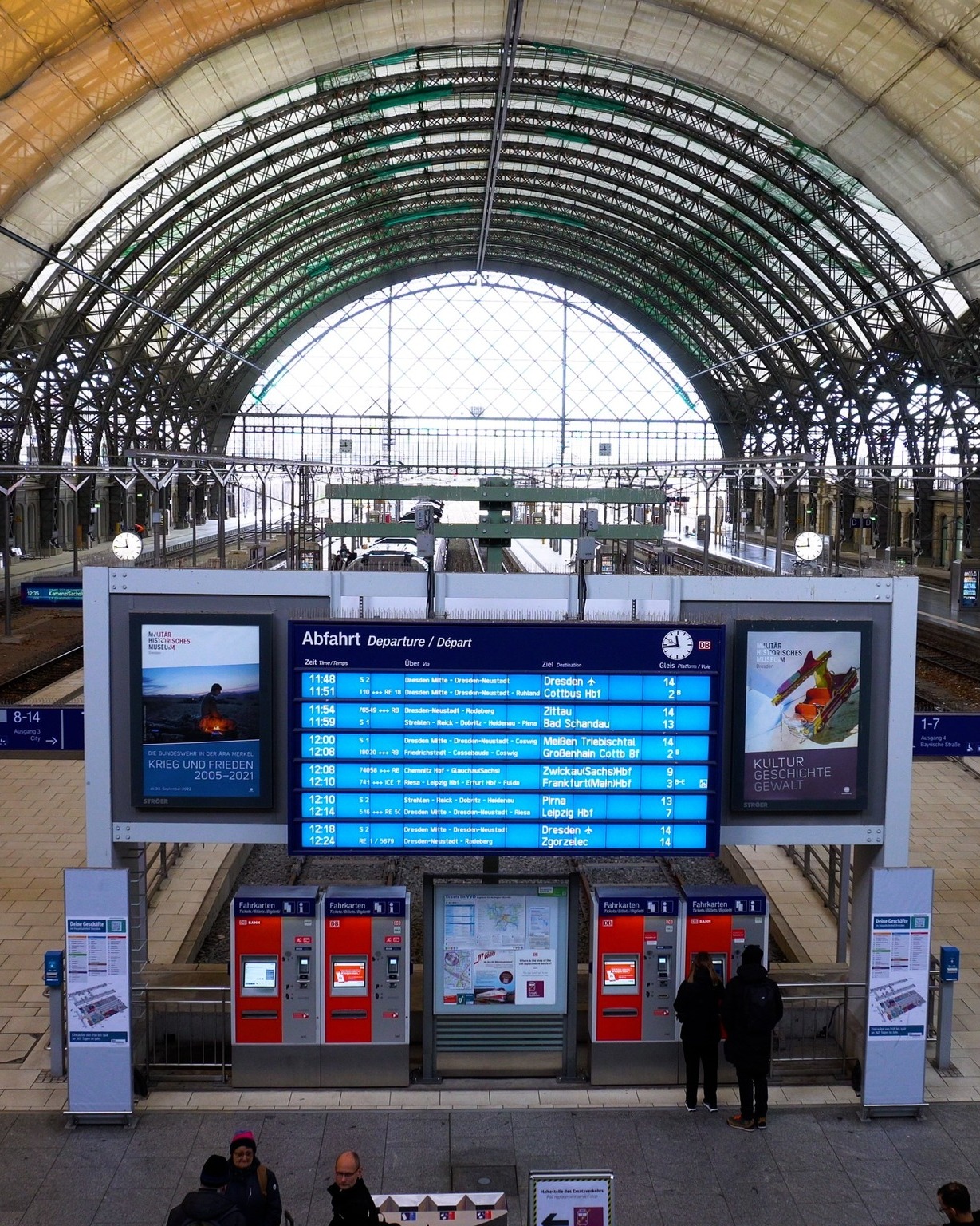 Dieser Alternativtext wurde KI-generiert: Ein Innenraum mit hoher, gewölbter Glasdecke. Im Vordergrund ein Zuganzeiger und Fahrkartenschalter.