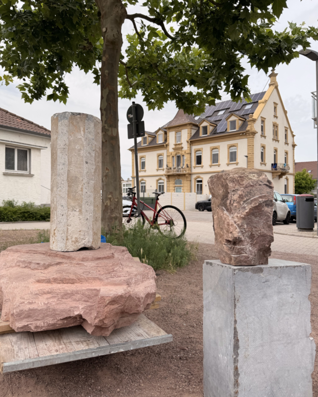 Zwei Steinobjekte auf Sockeln im Freien: links ein zylindrischer Stein auf einem roten Felsblock, rechts ein unregelmäßiger Stein auf einem grauen Betonsockel. Im Hintergrund ein Baum, ein rotes Fahrrad und ein gelbes Wohnhaus mit Solaranlage. (KI-Alternativtext)