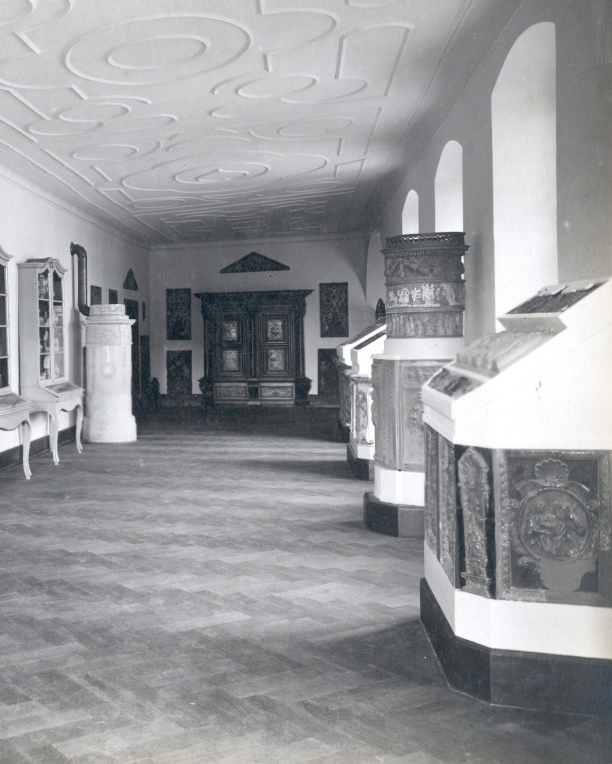 Dieser Alternativtext wurde KI-generiert: Schwarz-weißes Foto einer langen, leeren Halle mit dekorativer Stuckdecke. Entlang der Wände stehen Möbelstücke und Ausstellungsstücke.