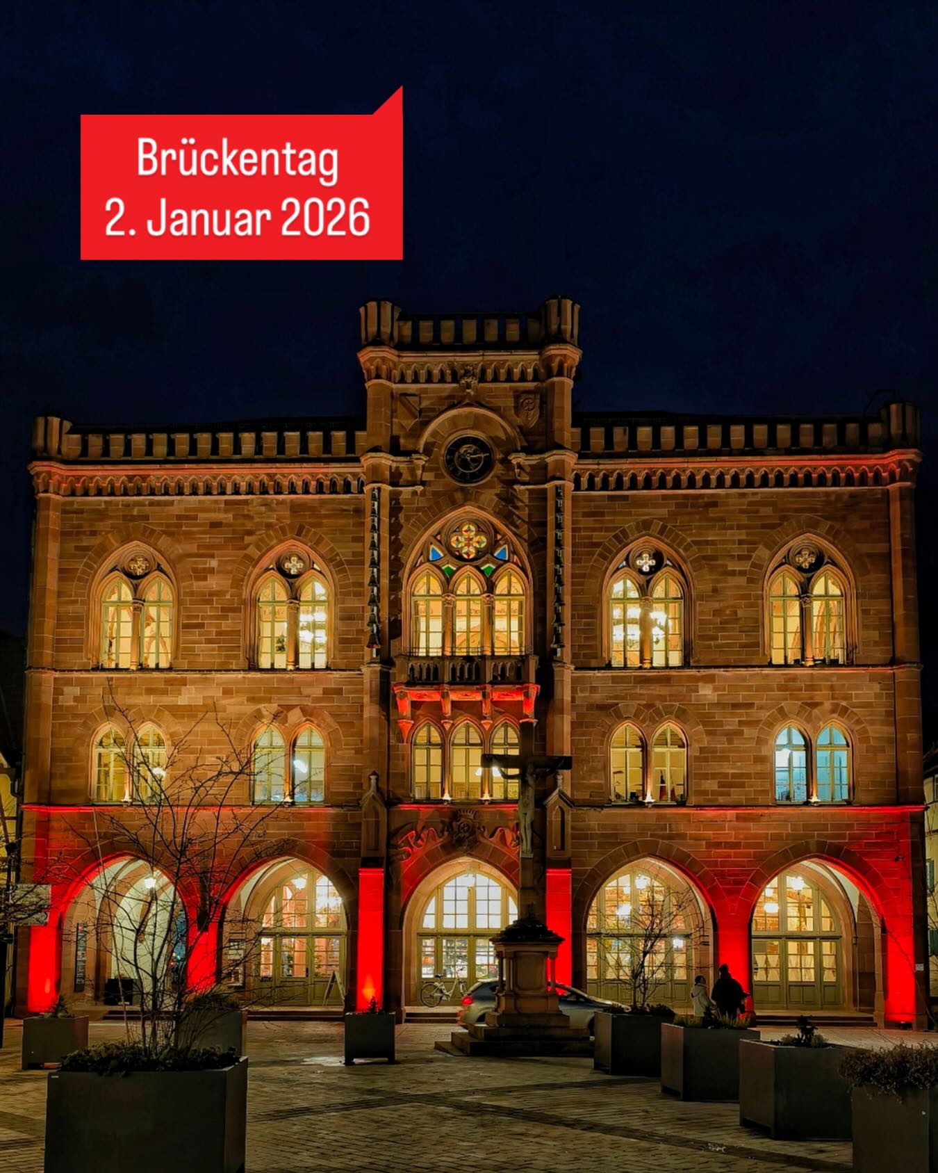 Dieser Alternativtext wurde KI-generiert: Ein beleuchtetes Gebäude mit vielen Fenstern und Bögen steht in der Nacht. Ein roter Hinweis mit Datumsangabe ist im Bild zu sehen.