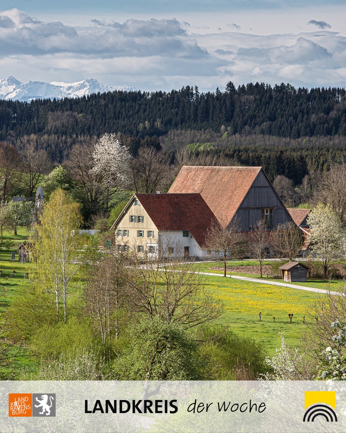 Dieser Alternativtext wurde KI-generiert: Eine ländliche Szene mit einem Bauernhaus und einem Feld im Vordergrund. Im Hintergrund sind bewaldete Hügel und schneebedeckte Berggipfel zu sehen.