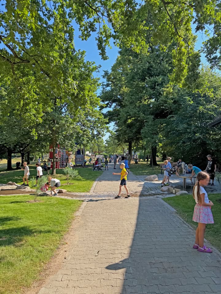 Ein sonniger Park mit einem gepflasterten Weg, der sich zwischen hohen Bäumen in die Ferne zieht. Eine Person in einem gelben Oberteil läuft über den Weg, während rechts eine Person in einem hellen Kleid steht und links andere Personen in einer Mulde spielen. Im Hintergrund sind Spielgeräte und eine Gruppe an einem Tisch zu sehen. (KI-Alternativtext)
