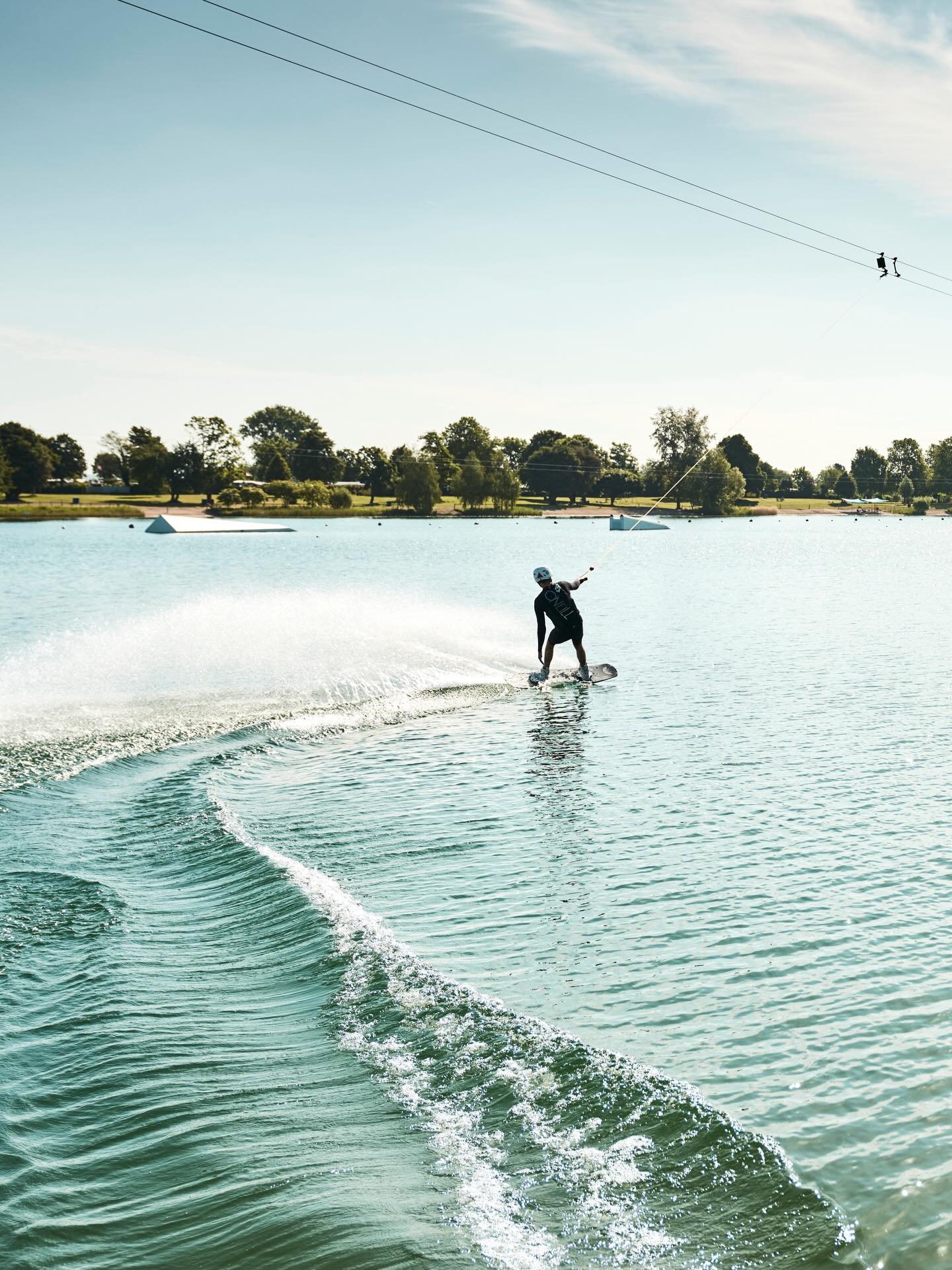 Eine Person in Neoprenanzug und Helm fährt auf einem Wakeboard über das Wasser und hält an einer Leine, die zu einem Seilzugsystem führt. Im Hintergrund sind Bäume und Rampen im Wasser zu sehen. (KI-Alternativtext)