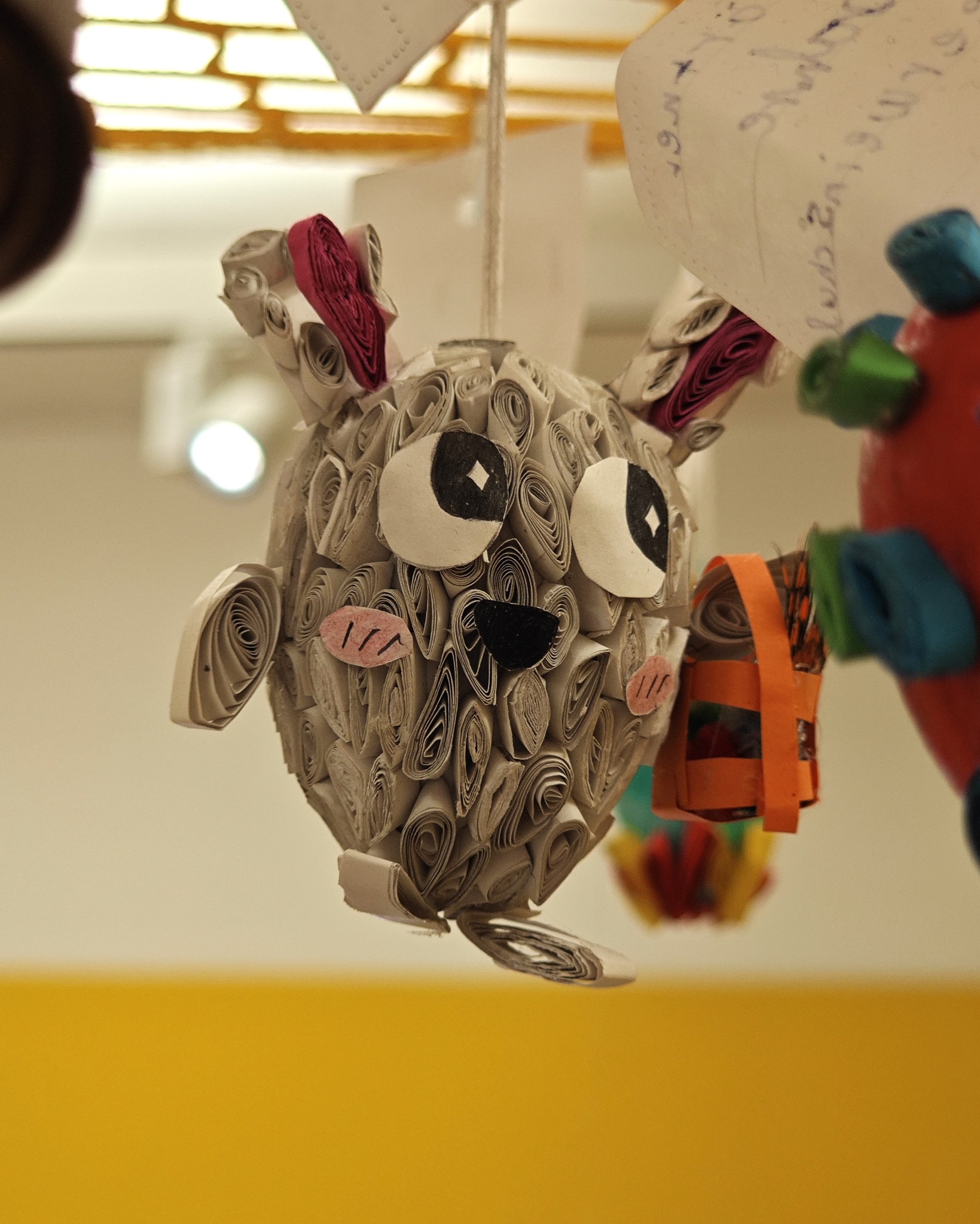 Dieser Alternativtext wurde KI-generiert: Ein mobiles Kunstwerk hängt von der Decke. Es zeigt eine kreative Figur aus gerolltem Papier, die an einen Tierkopf erinnert.
