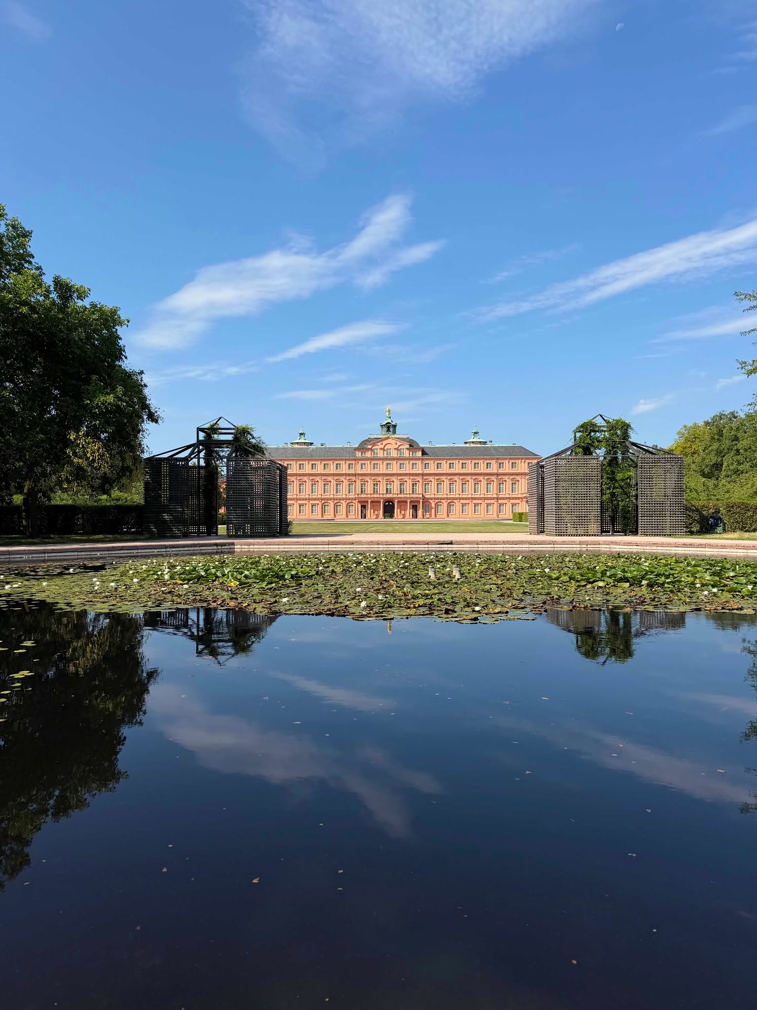 Ein Teich mit Seerosen im Vordergrund spiegelt den blauen Himmel. Im Hintergrund steht ein großes, rosa Schloss mit mehreren Fenstern und Türmen. Zwei dunkle Holzkonstruktionen flankieren den Blick auf das Gebäude. (KI-Alternativtext)