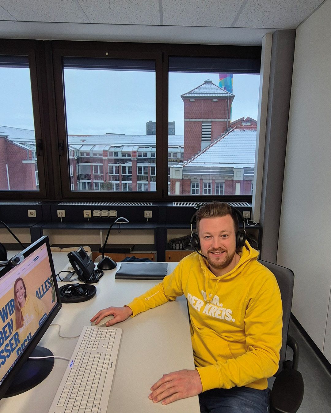 Dieser Alternativtext wurde KI-generiert: Ein Innenraum mit einer Person am Schreibtisch vor einem Fenster. Durch das Fenster sind Gebäude und ein grauer Himmel zu sehen.