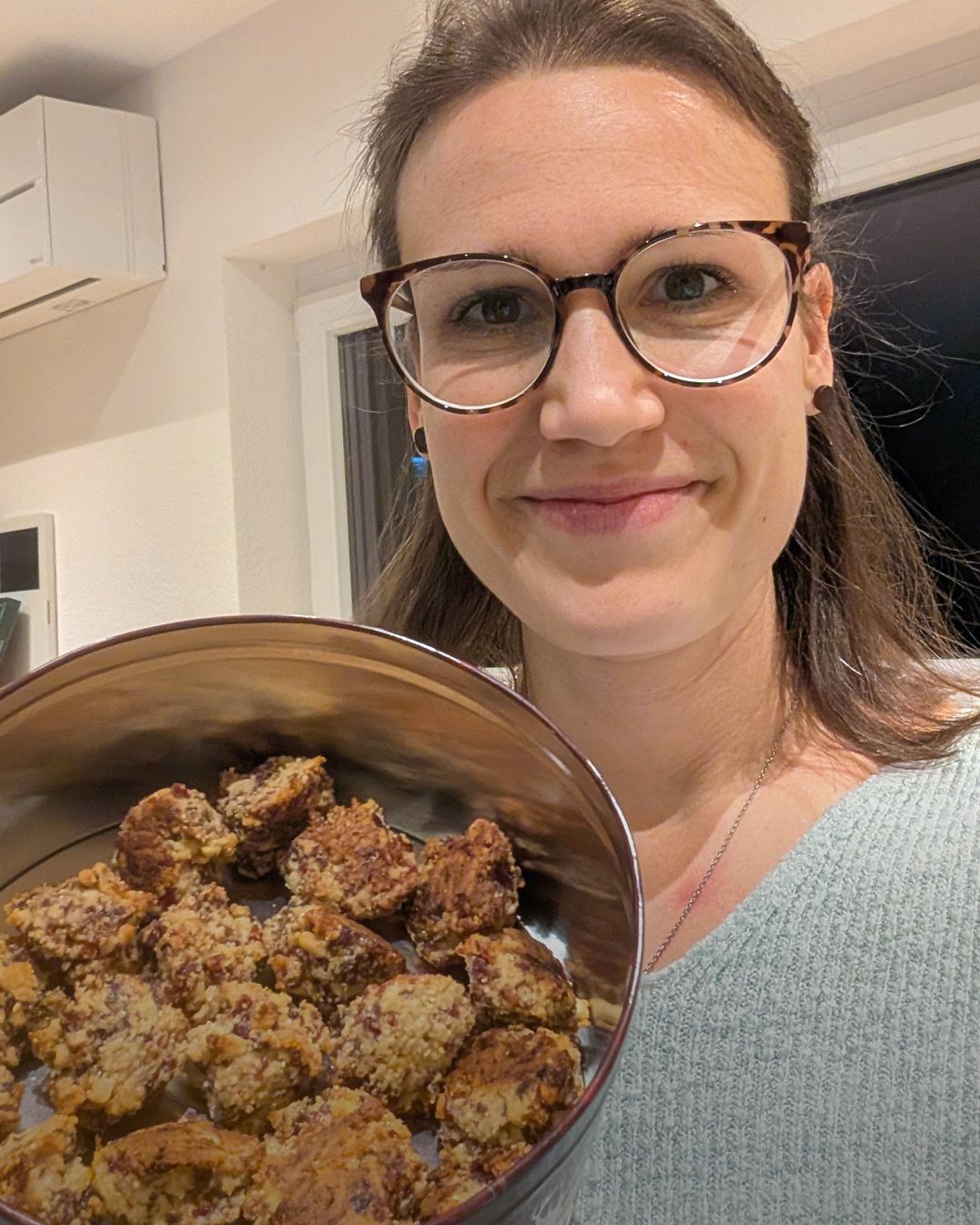 Dieser Alternativtext wurde KI-generiert: Ein Porträt von einer Person mit Brille, die ein Schüssel mit Essen hält. Im Hintergrund ist ein Raum mit Möbeln zu erkennen.