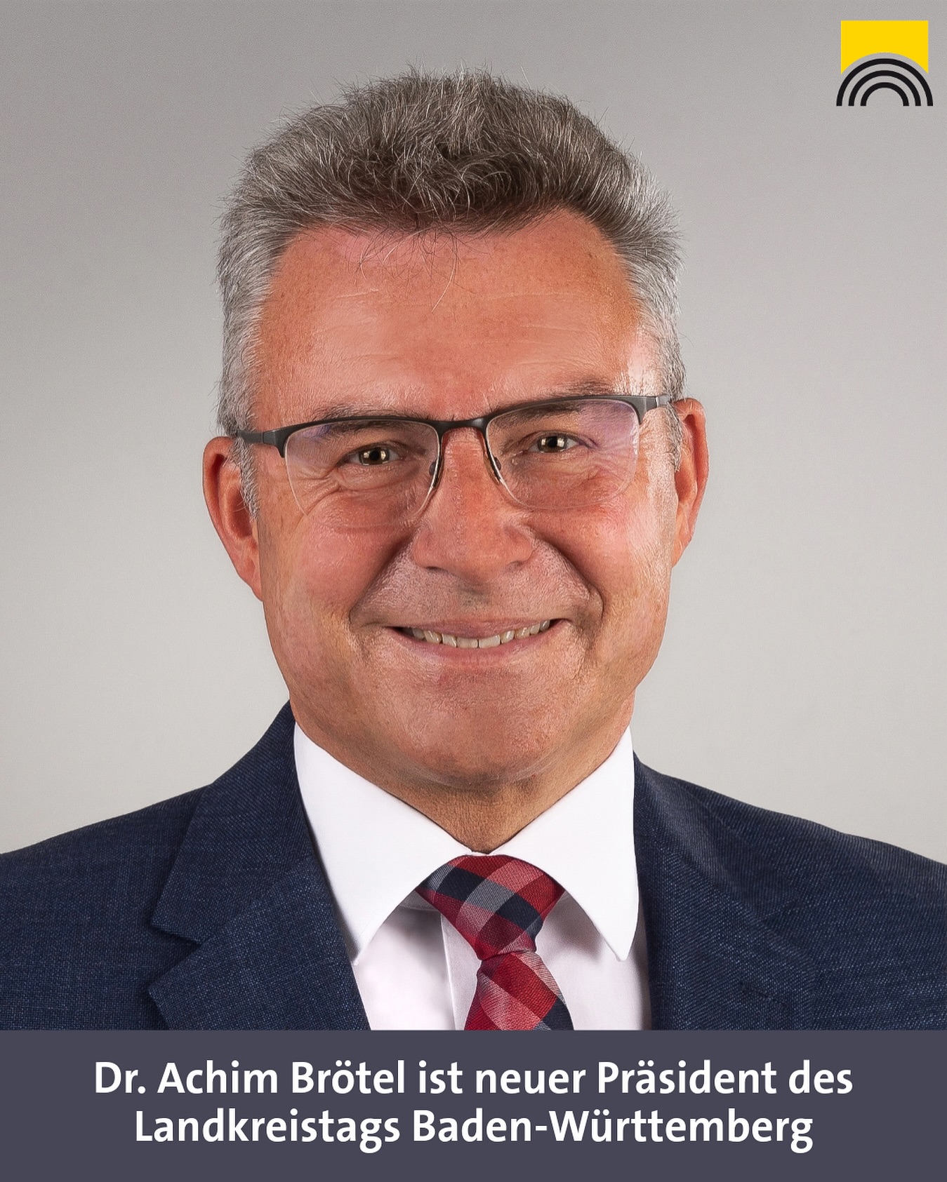 Dieser Alternativtext wurde KI-generiert: Porträt einer Person mit Brille und Anzug, vor einem hellgrauen Hintergrund. Im Bild ist ein farbiger Balken im oberen rechten Eck zu sehen.