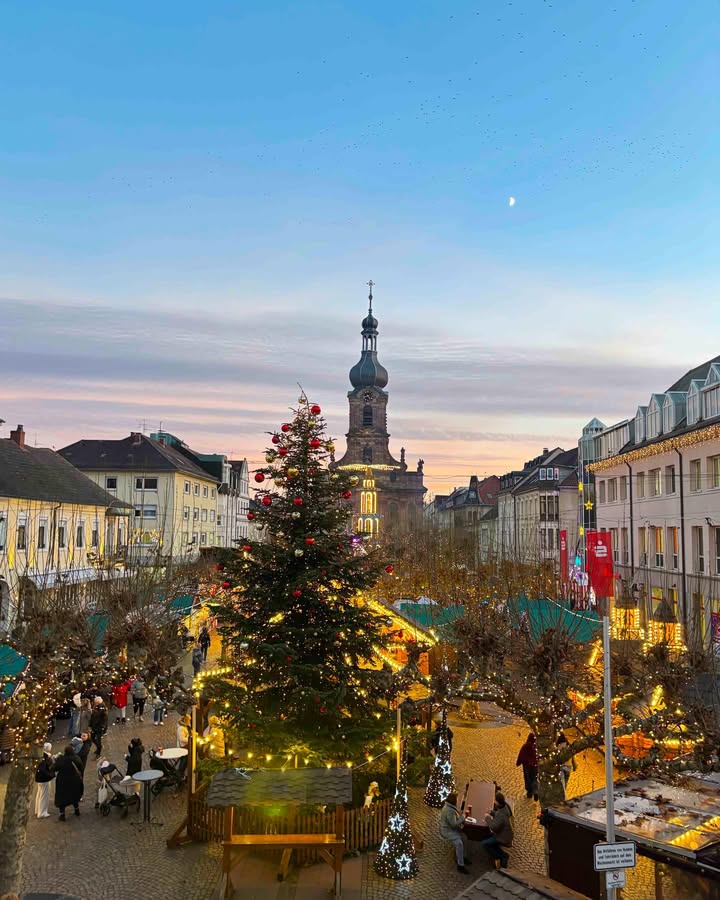 Dieser Alternativtext wurde KI-generiert: Ein Stadtplatz mit Weihnachtsmarkt, dekoriert mit Lichtern und einem Weihnachtsbaum. Im Hintergrund ist ein hoher Kirchturm unter einem rosafarbenen Himmel zu sehen.