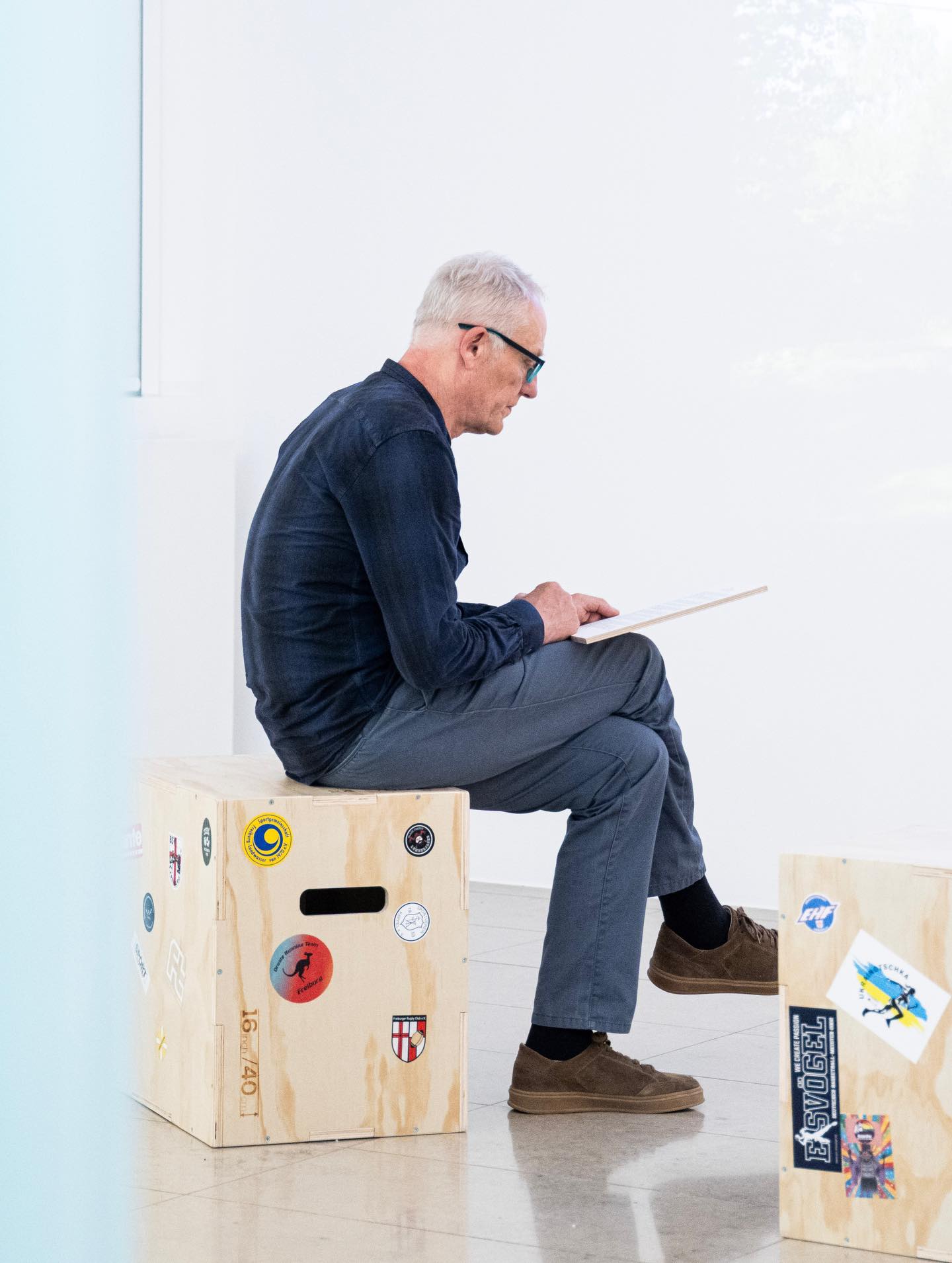 Dieser Alternativtext wurde KI-generiert: Eine Person sitzt auf einem Holzstück und schreibt auf ein Blatt Papier. Im Hintergrund sind weitere Holzstücke und eine helle Umgebung zu sehen.