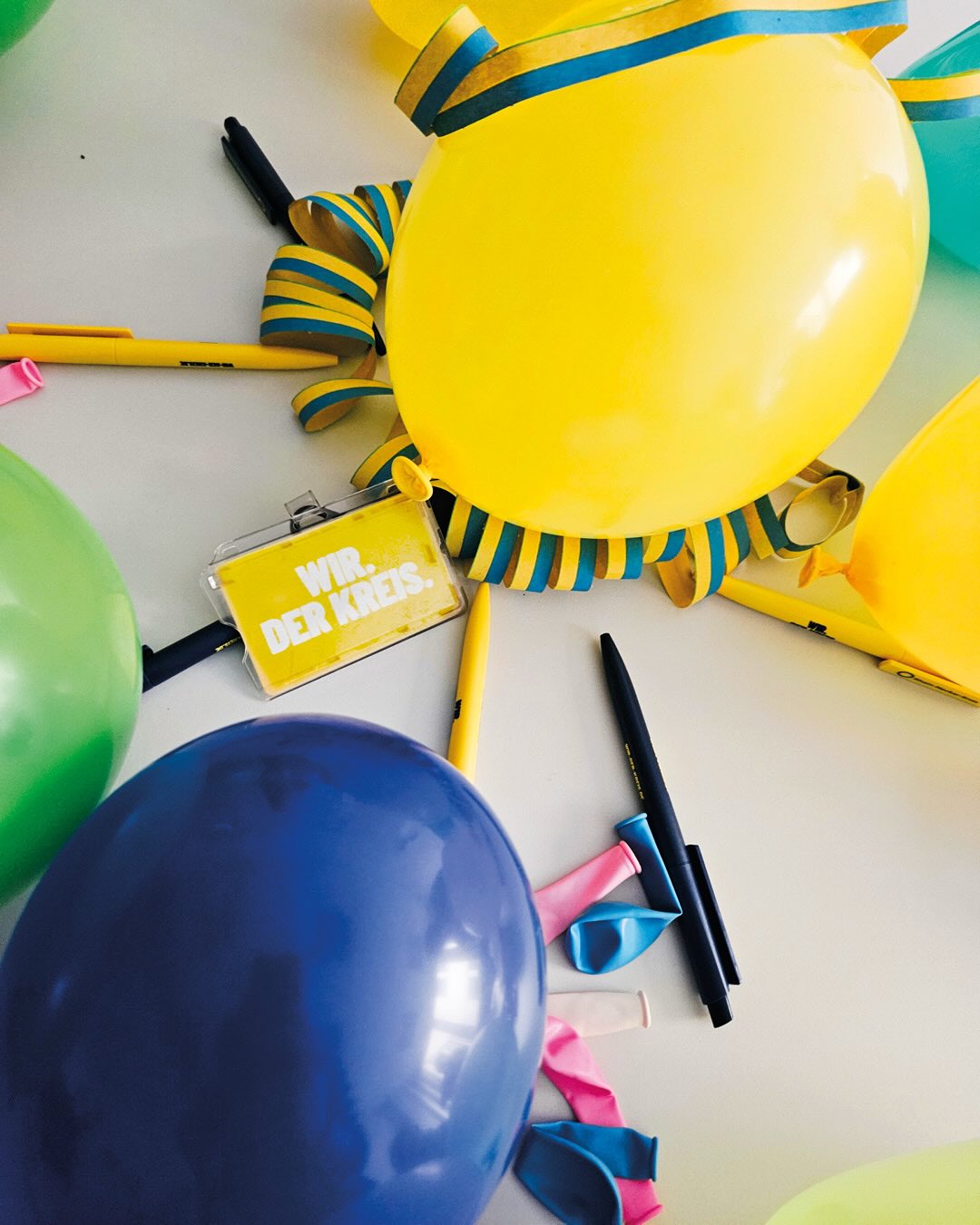 Dieser Alternativtext wurde KI-generiert: Farbenfrohe Dekorationen liegen auf einer hellen Oberfläche. Luftballons, Girlanden und Schreibutensilien sind zu sehen.