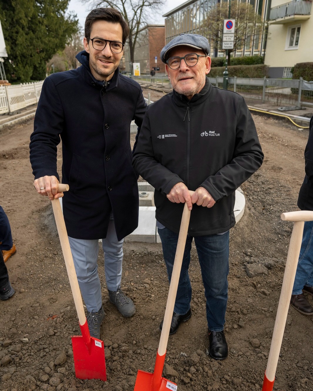 Dieser Alternativtext wurde KI-generiert: Zwei Personen im Freien stehen nebeneinander und halten Spaten. Im Hintergrund sind Gebäude und eine Baustelle zu sehen.