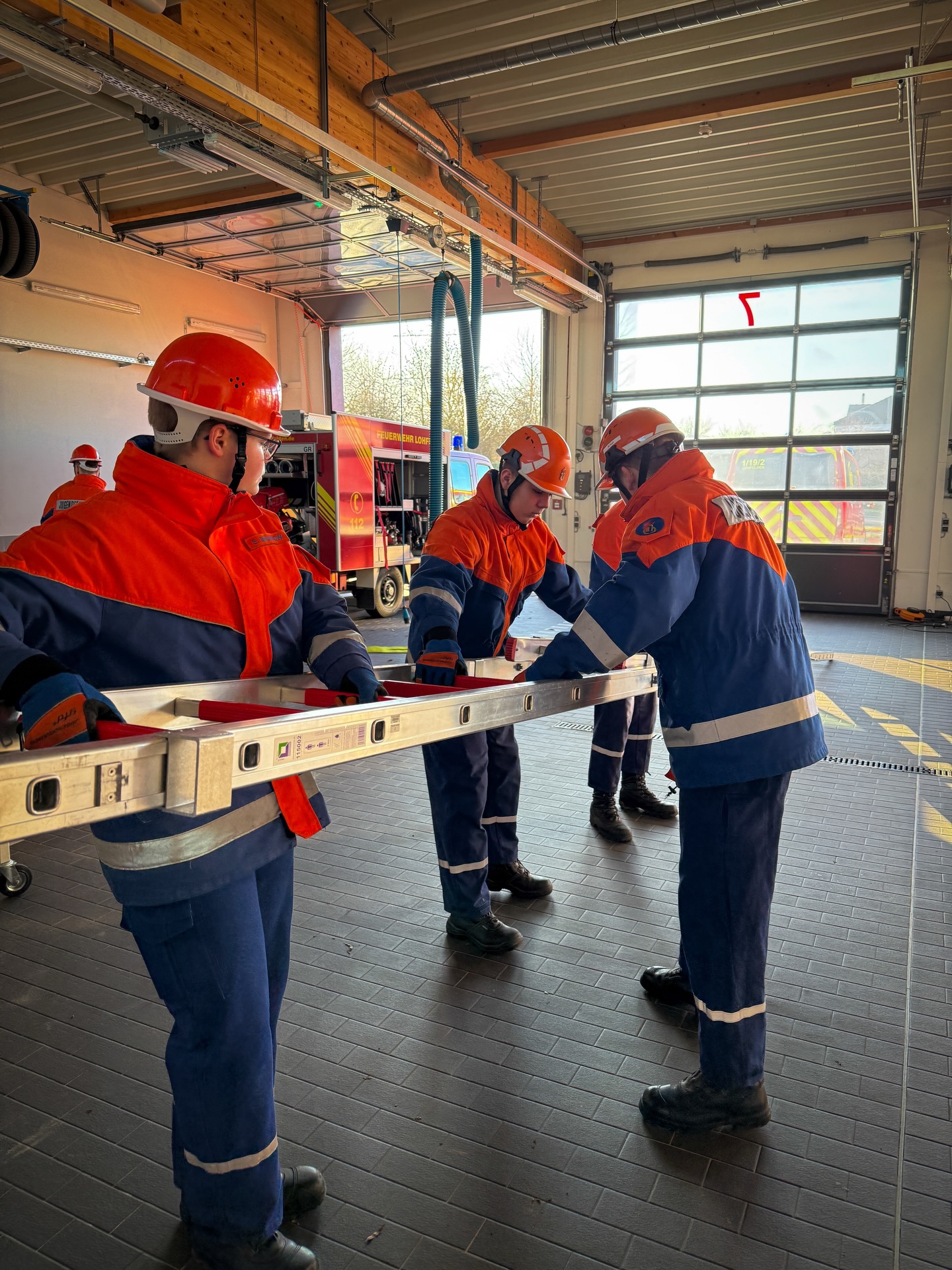 Drei Personen in orangefarbenen und blauen Feuerwehruniformen mit Helmen halten gemeinsam eine lange, silberne Leiter waagerecht. Im Hintergrund der Halle steht ein rotes Fahrzeug mit der Aufschrift „FEUERWEHR“ und eine große Toreinfahrt mit der roten Nummer 7. (KI-Alternativtext)