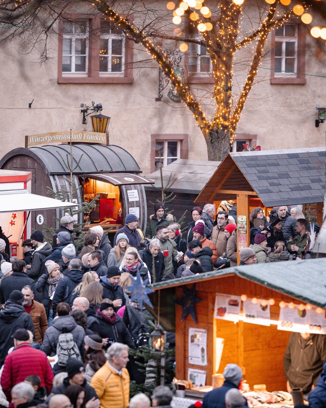 Dieser Alternativtext wurde KI-generiert: Ein belebter Marktplatz mit mehreren Ständen und Lichtern. Menschen stehen und gehen zwischen den Ständen, die weihnachtliche Waren anbieten.
