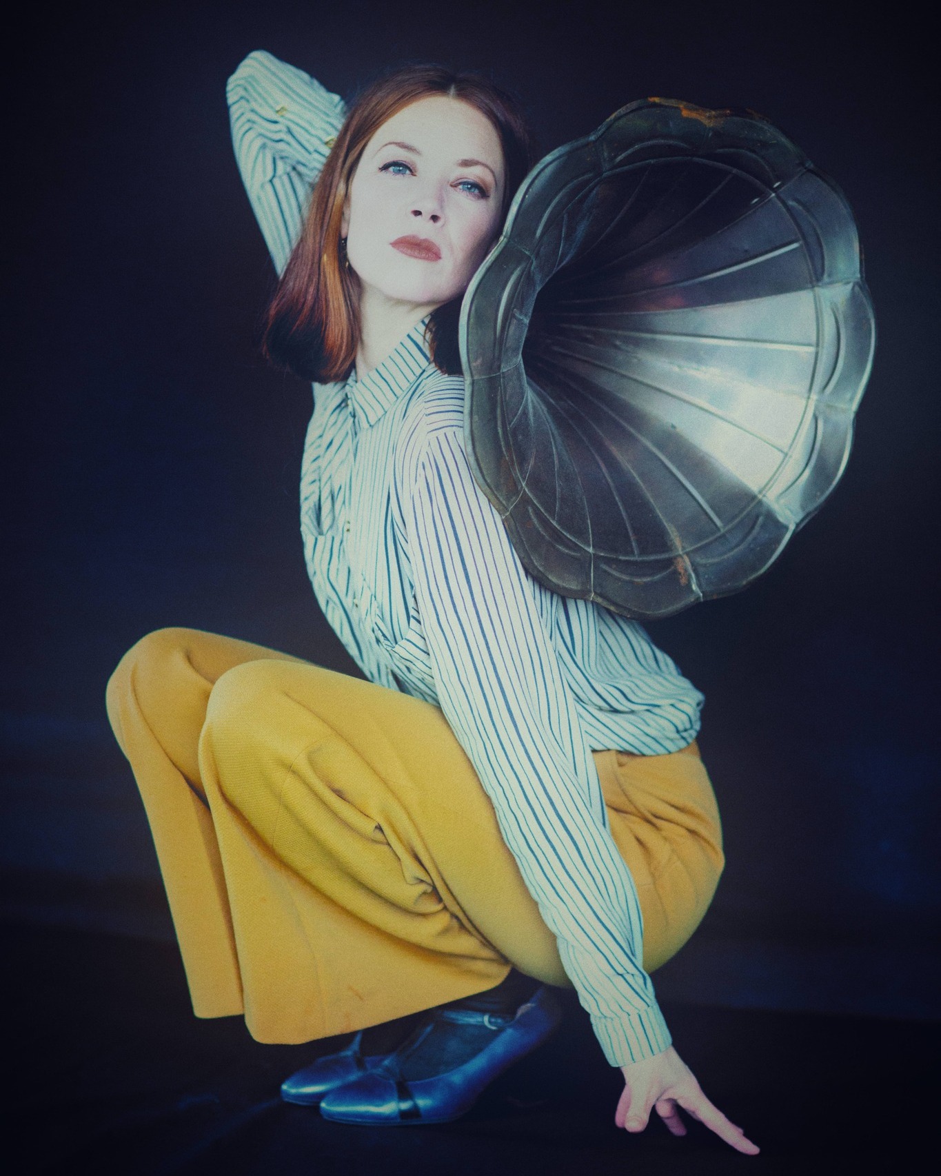 Dieser Alternativtext wurde KI-generiert: Eine Person in gestreifter Bluse und gelber Hose kauert und hält einen Schalltrichter vor das Ohr. Das Bild ist in einem markanten Farbton gehalten.