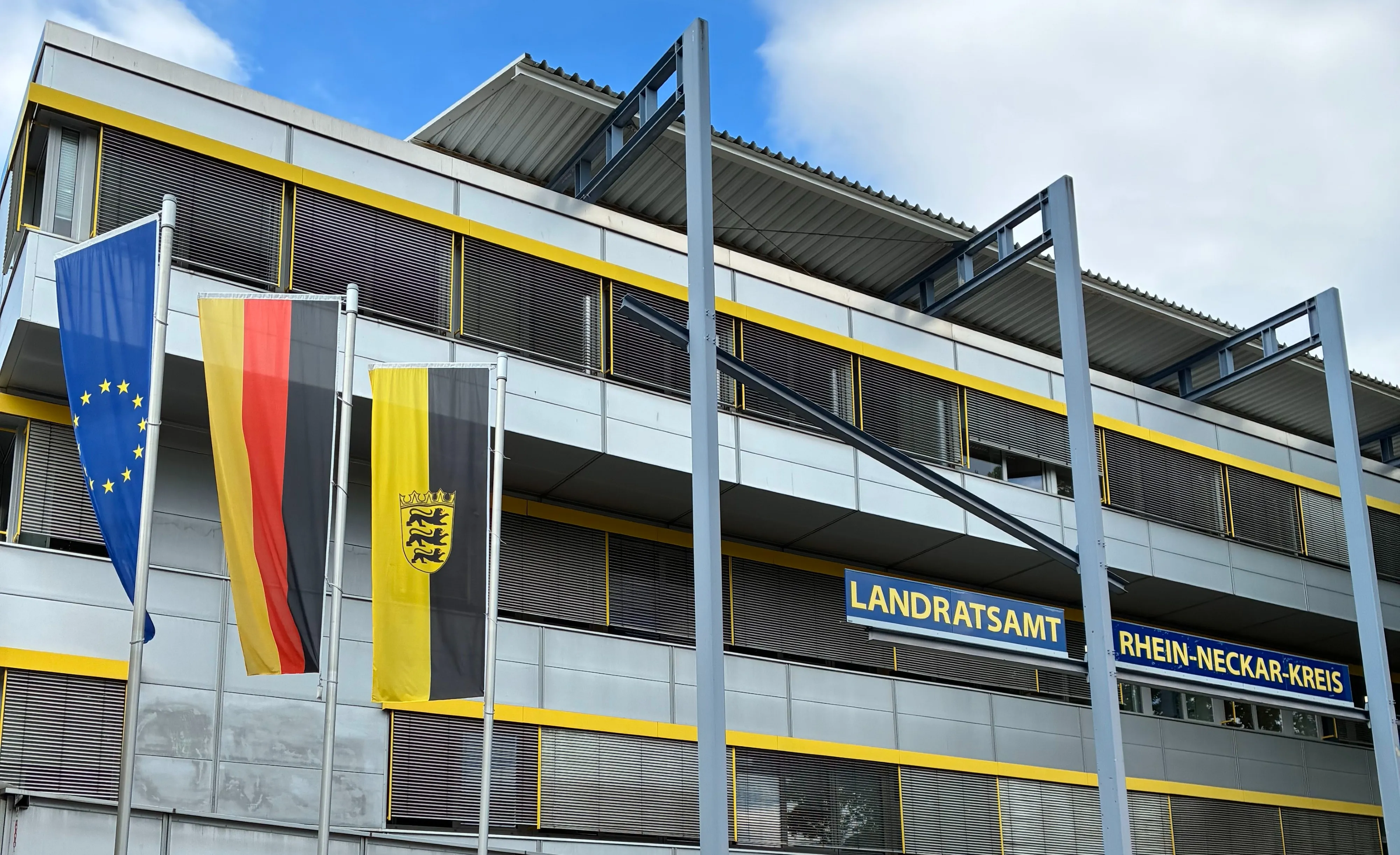 Fassade des Landratsamts Rhein-Neckar-Kreis mit drei Flaggen: der Europäischen Union, Deutschlands und des Rhein-Neckar-Kreises. Das Gebäude hat ein modernes Design mit gelben Akzenten und Jalousien. (KI-Alternativtext)