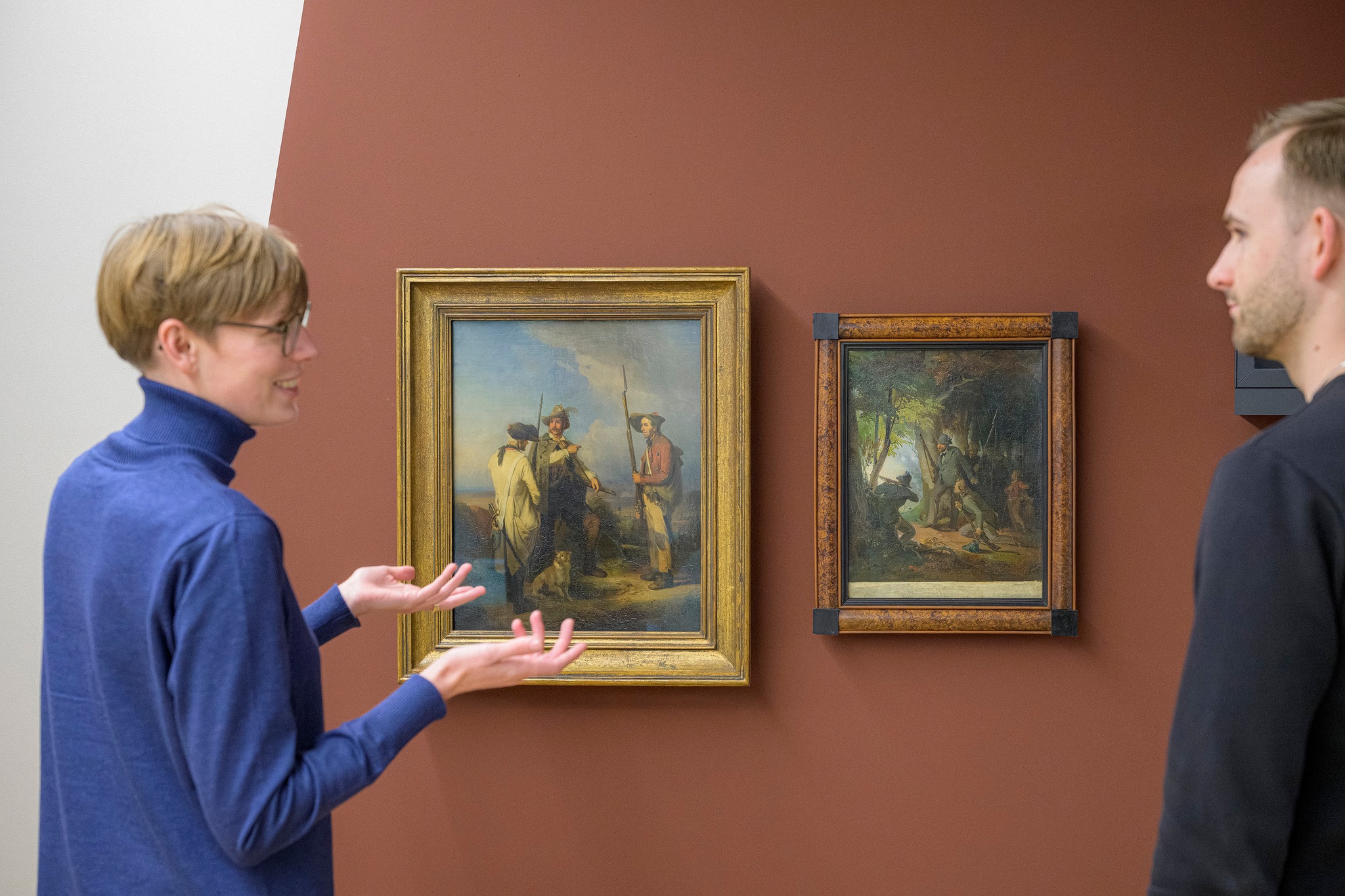 Eine Person in blauem Rollkragenpullover erklärt mit ausgestreckten Händen zwei Gemälde in goldenen Rahmen an einer braunen Wand, während eine zweite Person zuhört. Die Bilder zeigen historische Szenen mit mehreren Figuren, eine im Freien mit Hunden, die andere im Wald. (KI-Alternativtext)