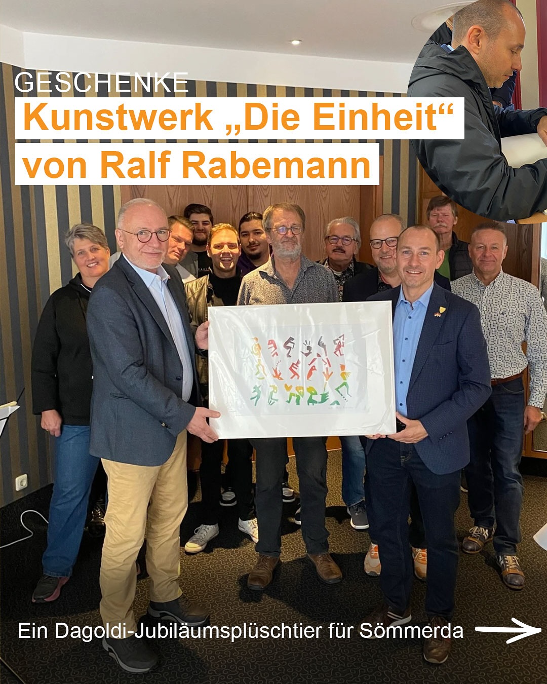 Dieser Alternativtext wurde KI-generiert: Eine Gruppe von Personen steht in einem Raum zusammen. Zwei Personen im Vordergrund halten ein großes, bemalte Blatt Papier.