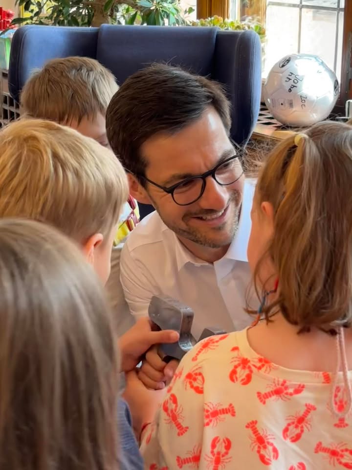 Ein Erwachsener mit Brille und weißem Hemd lächelt und hält gemeinsam mit einem Kind einen grauen Spielzeughammer. Im Vordergrund steht ein Kind mit einem hellen Oberteil, das ein rotes Muster aufweist. Im Hintergrund sind ein blaues Sofa und ein silberner Ballon mit Schrift zu erkennen. (KI-Alternativtext)