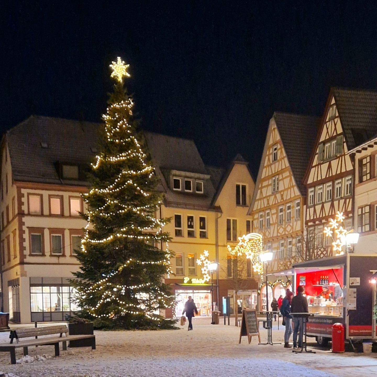 Dieser Alternativtext wurde KI-generiert: Ein beleuchteter Weihnachtsbaum steht auf einem Platz vor historischen Fachwerkhäusern. Im Hintergrund sind weitere Lichter und eine kleine Bewirtung zu sehen.