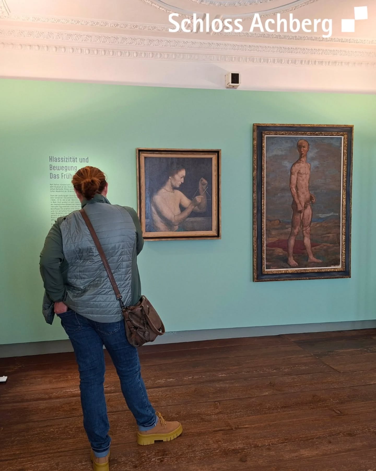 Dieser Alternativtext wurde KI-generiert: Ein Innenraum mit einer Person, die ein Gemälde betrachtet. Das Gemälde zeigt eine Figur und hängt an einer hellen Wand.