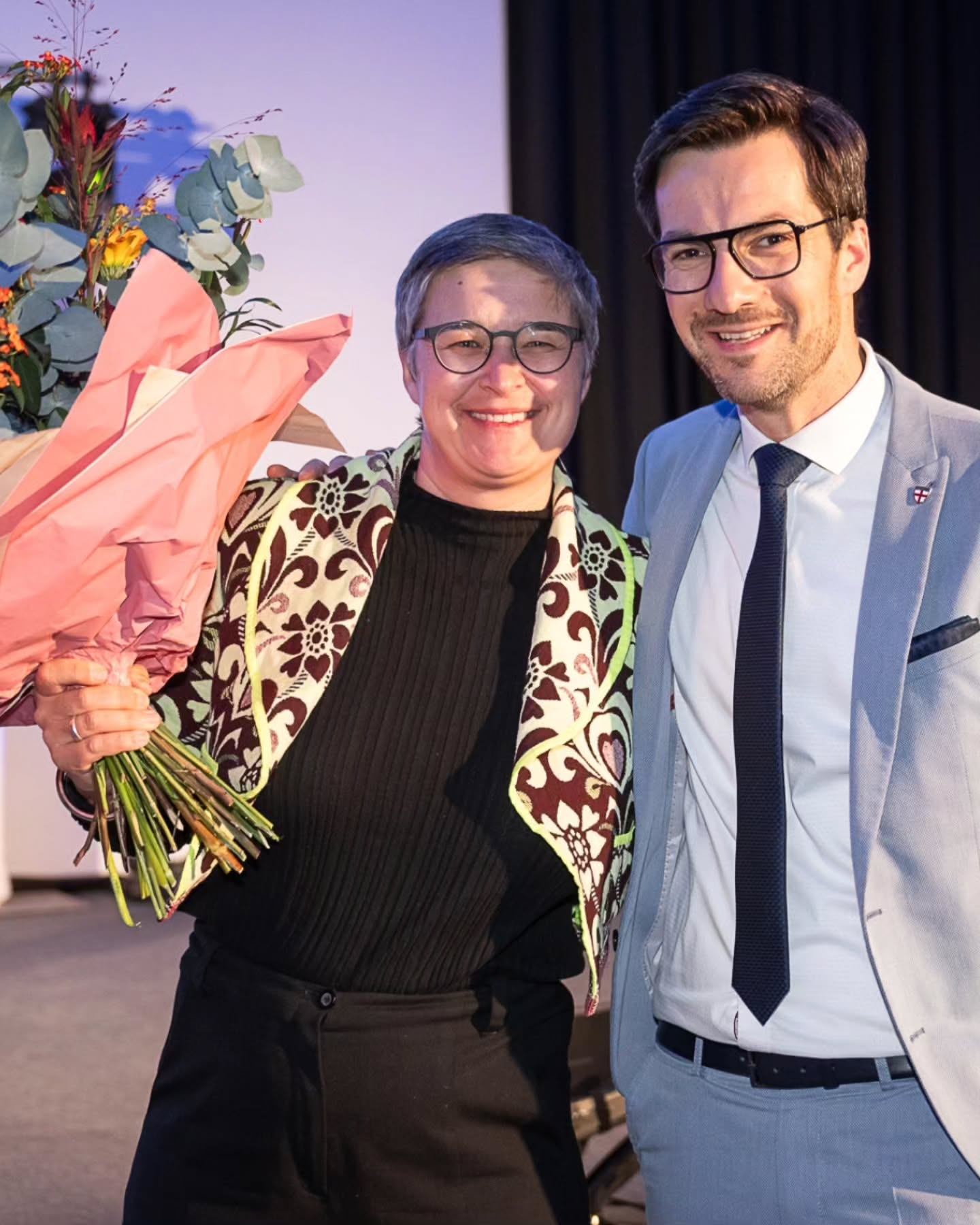 Dieser Alternativtext wurde KI-generiert: Zwei Personen stehen nebeneinander und lächeln in die Kamera. Sie halten Blumensträuße vor einem hellen Hintergrund.