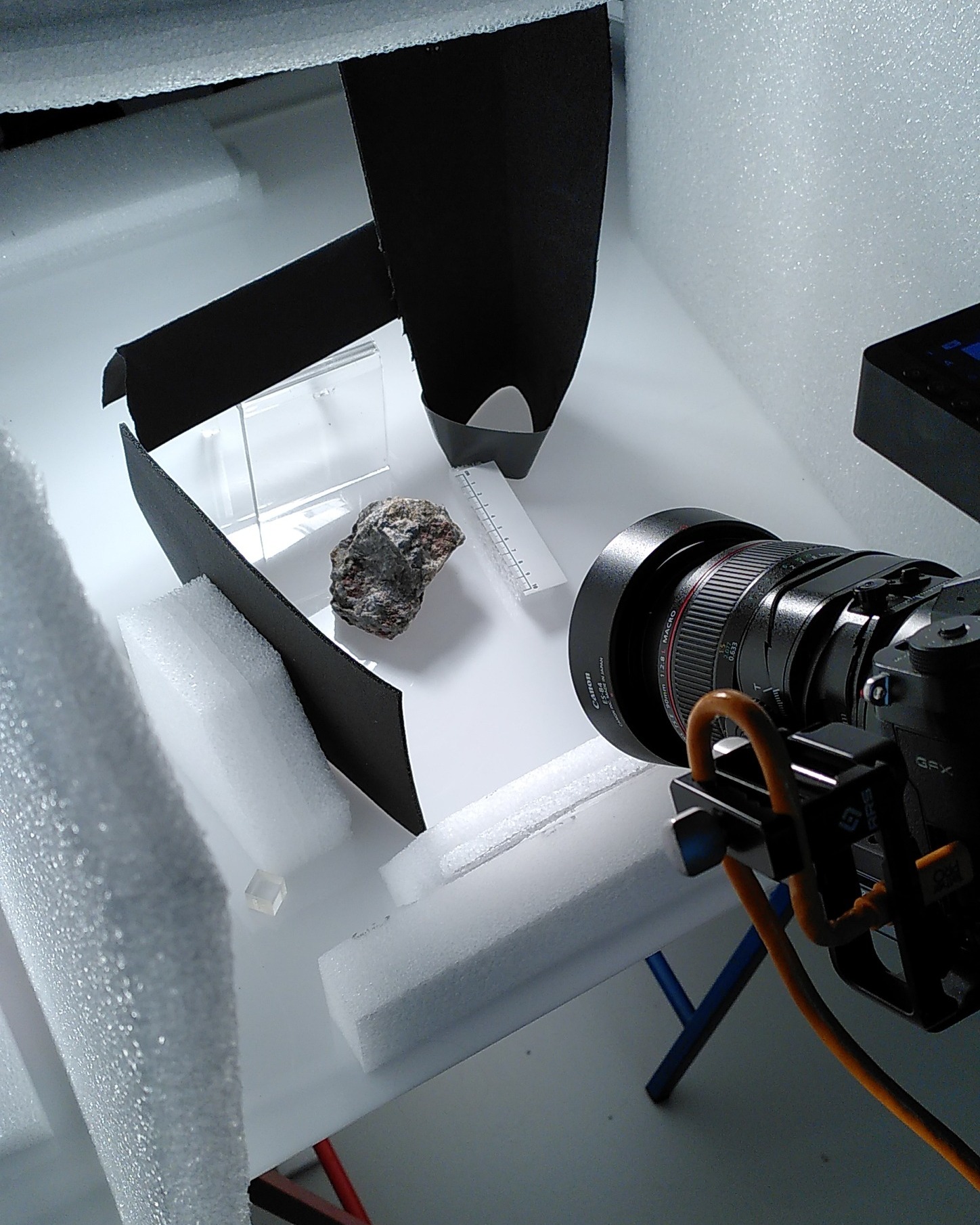 Dieser Alternativtext wurde KI-generiert: Ein Objekt wird von einer Kamera mit zusätzlichem Licht fotografiert. Das Arrangement befindet sich auf einer weißen, strukturierten Oberfläche.