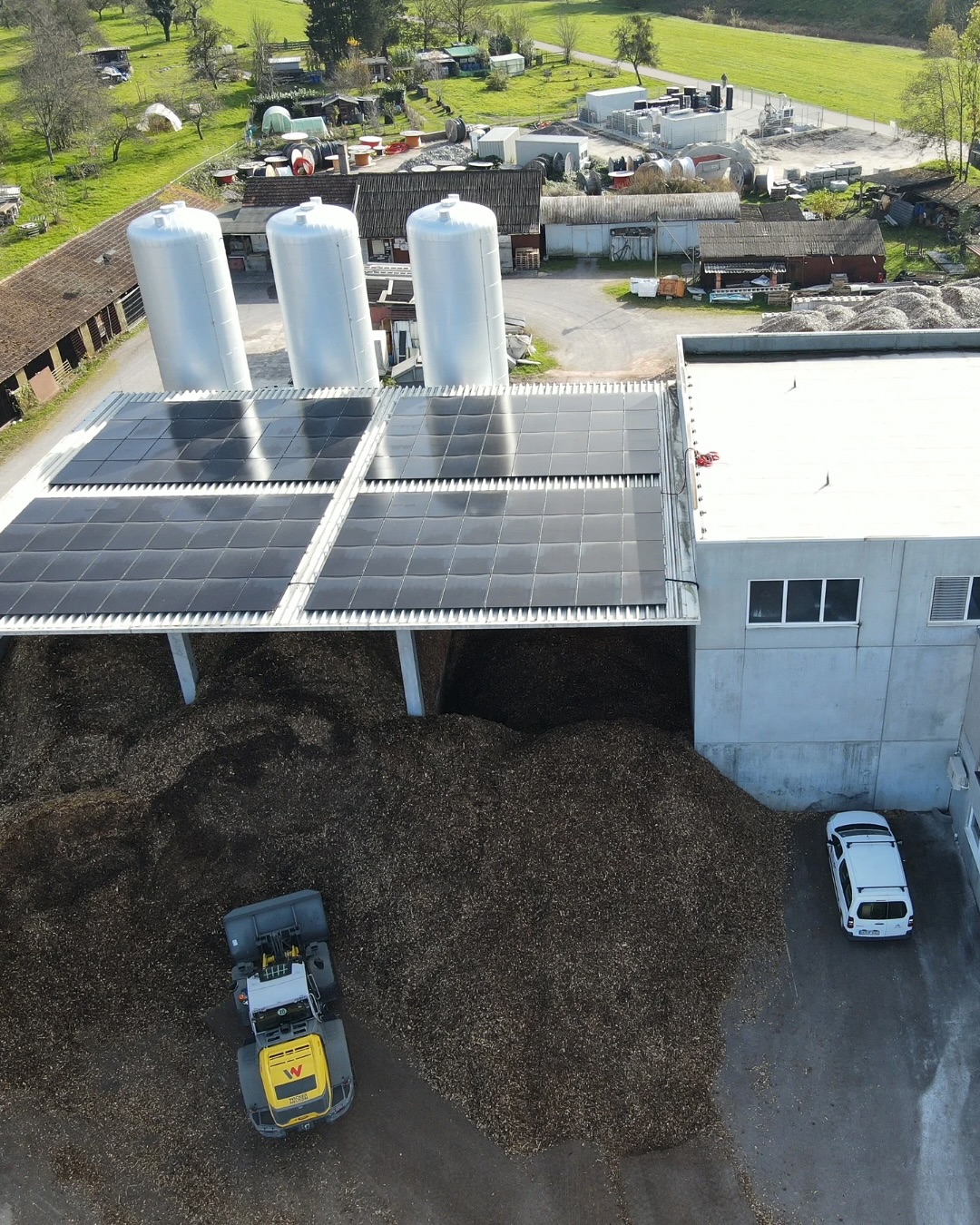 Eine Luftaufnahme von einem Gebäude mit Solarzellen auf dem Dach, großen Tanks und einem Haufen Material, neben dem ein Bagger steht.