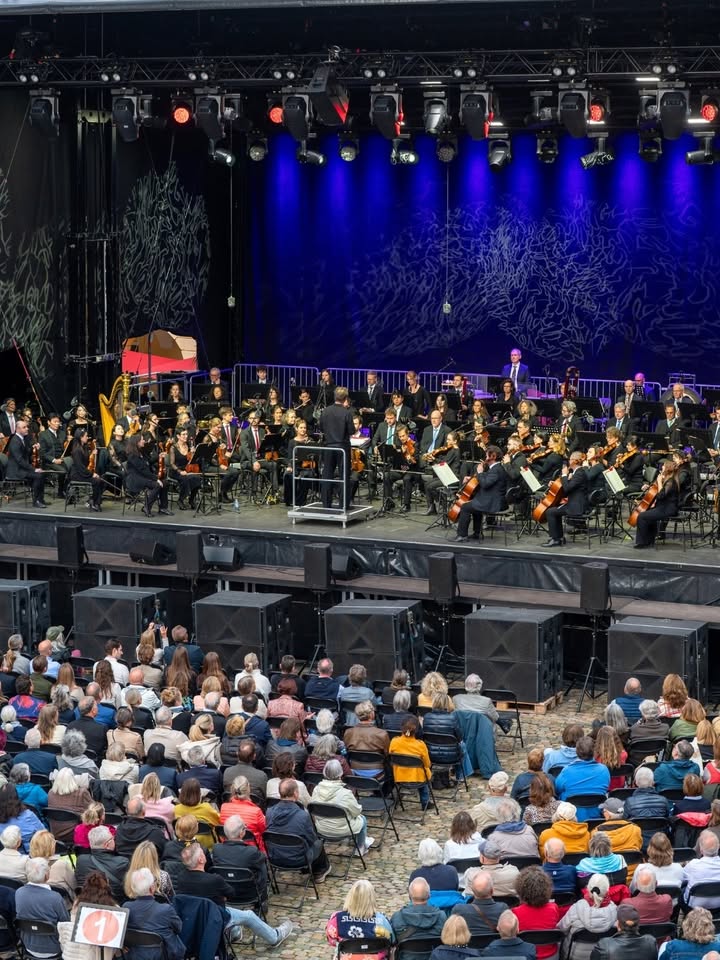 Ein Orchester auf einer Bühne mit blauer Beleuchtung und einem Dirigenten im Vordergrund, vor einem Publikum, das auf Stühlen sitzt und zuschaut. (KI-Alternativtext)