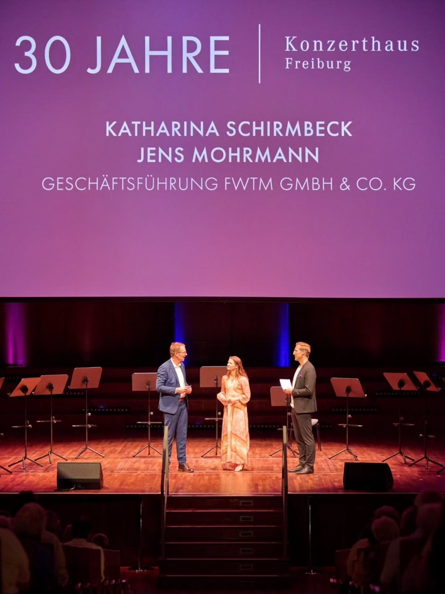 Drei Personen stehen auf einer Bühne vor einem großen violetten Bildschirm, auf dem steht: 30 JAHRE | Konzerthaus Freiburg, KATHARINA SCHIRMBECK, JENS MOHRMANN, GESCHÄFTSFÜHRUNG FW TM GMBH & CO. KG. Hinter den Personen stehen Notenständer, im Vordergrund sind die Köpfe des Publikums zu sehen. (KI-Alternativtext)