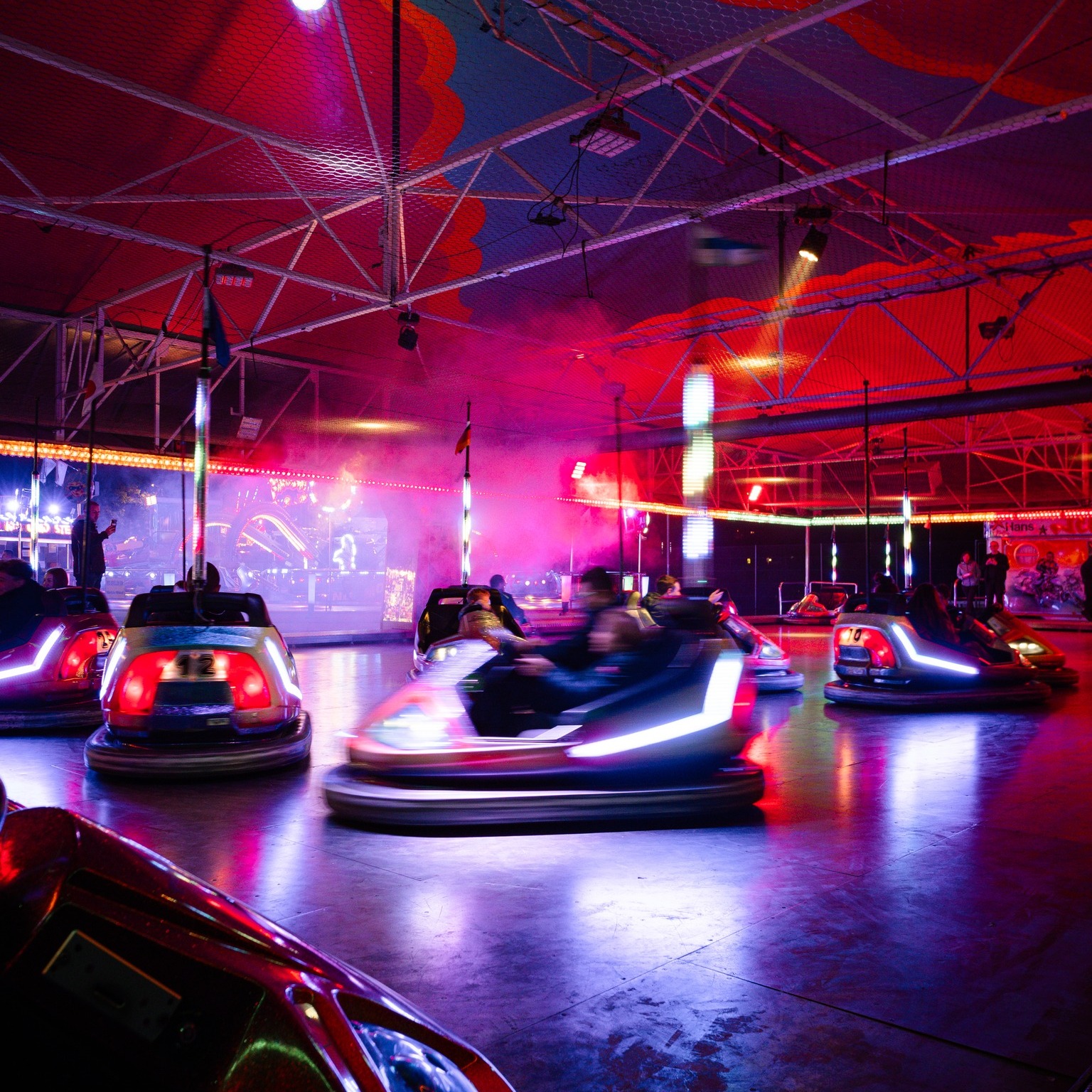 Dieser Alternativtext wurde KI-generiert: Ein Innenraum mit einer überdachten Struktur, der in intensive Rottöne und Violett getaucht ist. Im Vordergrund sind bewegte Autos zu sehen, vermutlich in einer Art Fahrgeschäft.