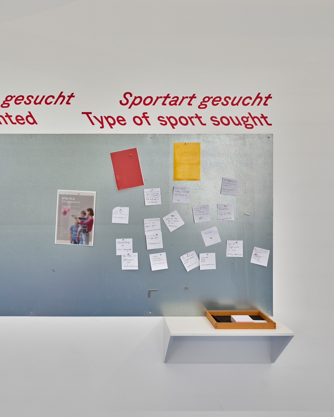 Rote Schrift auf einer weißen Wand lautet "Sportart gesucht Type of sport sought". Darunter befindet sich ein graues Pinnwand-Board, auf dem ein rotes und ein gelbes Blatt Papier, ein Flyer für "SPIELFELD Schnupperkurse" sowie zahlreiche kleine weiße Notizen mit handschriftlichen Texten befestigt sind. Unterhalb der Pinnwand steht ein weißes Regal mit einem Holztablett. (KI-Alternativtext)