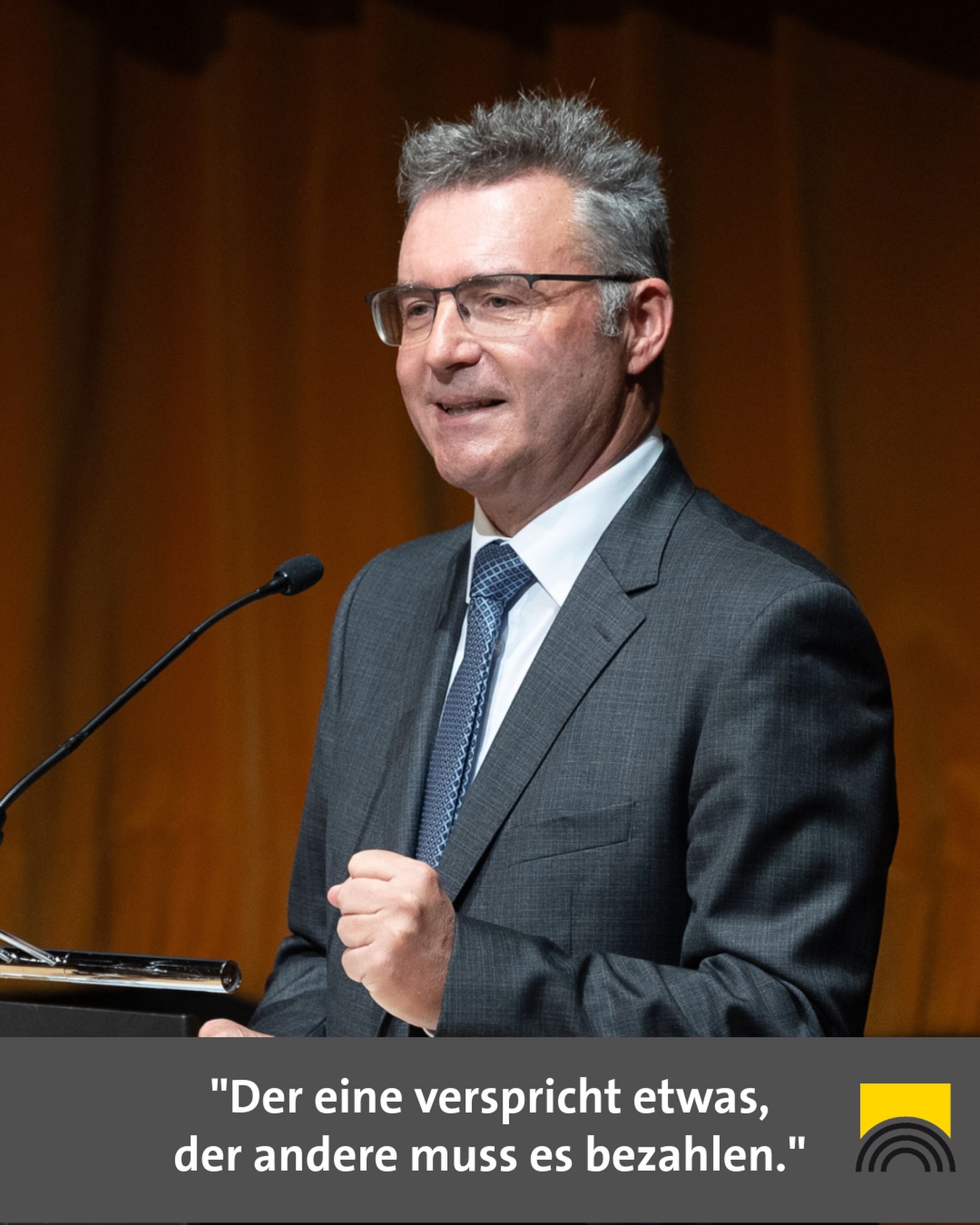 Dieser Alternativtext wurde KI-generiert: Eine Person mit Brille spricht in ein Mikrofon auf einer Bühne. Ein Zitat ist am unteren Bildrand zu sehen.