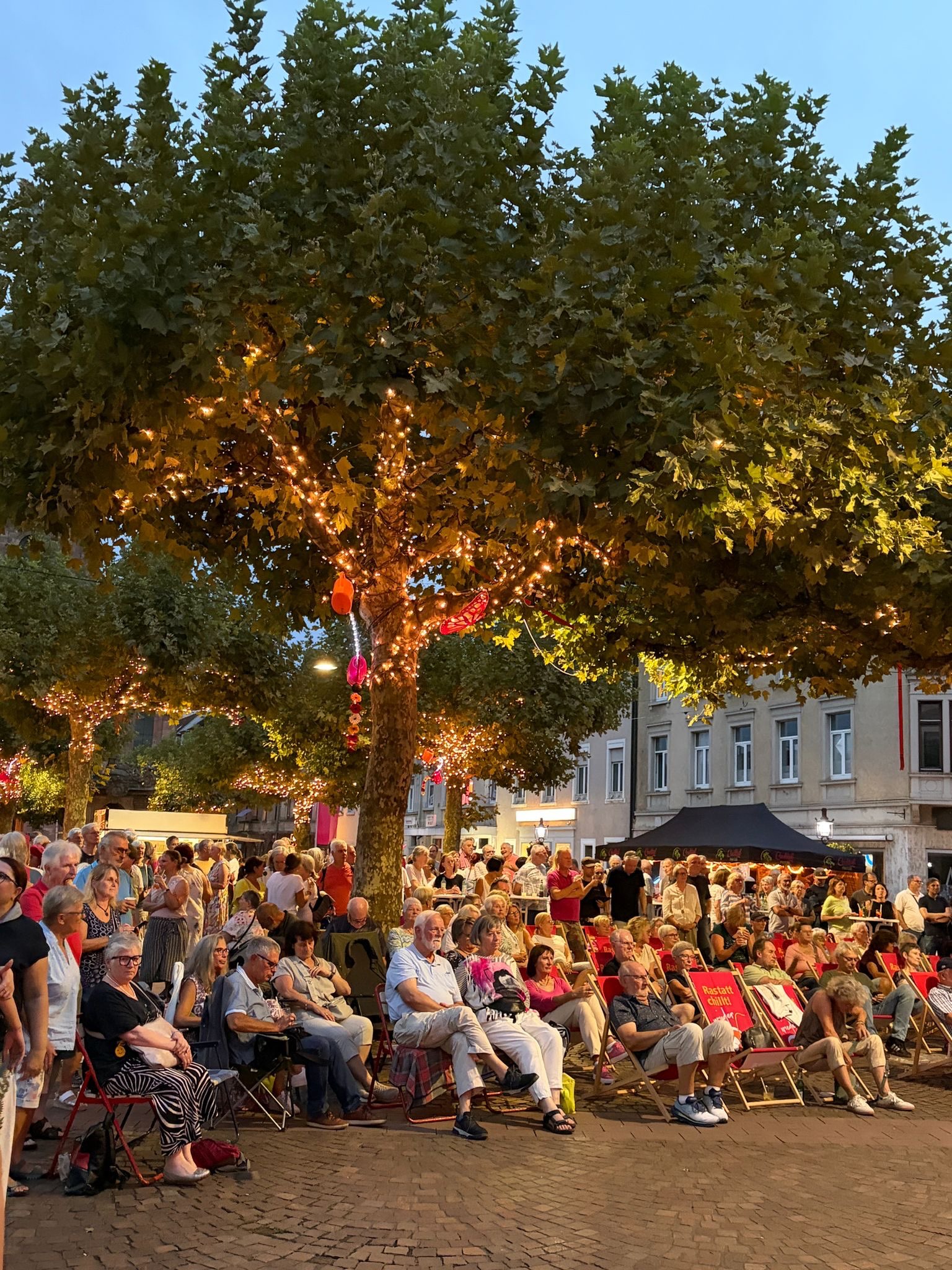 Viele Personen sitzen auf roten Klappstühlen auf einem gepflasterten Platz oder stehen im Hintergrund. Ein großer Baum mit Lichterketten und bunten Dekorationen steht im Vordergrund. Auf der Rückenlehne eines Stuhls ist der Text „Rastatt chillt!“ zu lesen. (KI-Alternativtext)