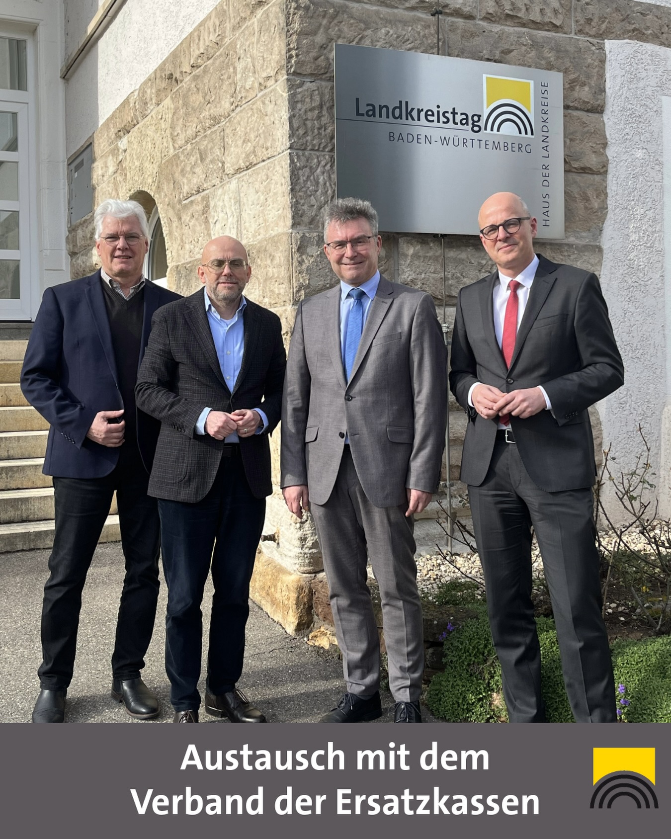 Dieser Alternativtext wurde KI-generiert: Vier Personen in Anzügen stehen vor einem Gebäude mit einem Banner. Darunter befindet sich ein Text und ein Logo.