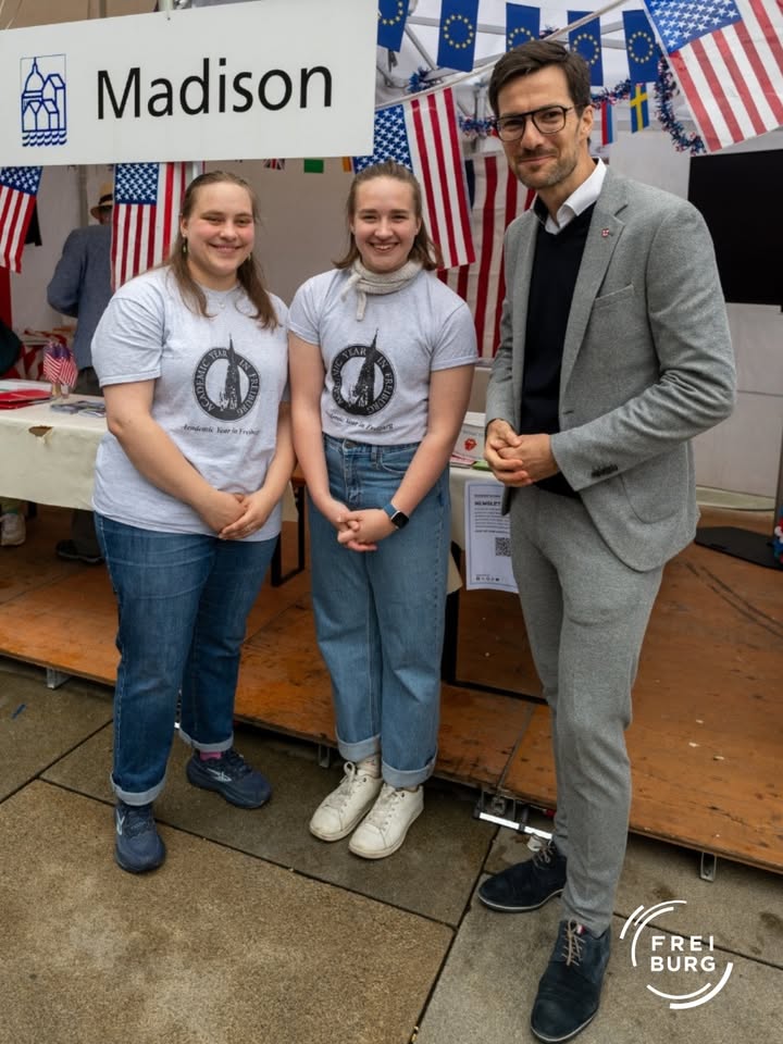Drei Personen stehen vor einem Stand mit dem Schild „Madison“ und US-Flaggen. Zwei tragen identische T-Shirts mit einem Rundlogo, die dritte Person trägt einen grauen Anzug. Im Hintergrund sind weitere Flaggen und ein Tisch mit Materialien zu sehen. (KI-Alternativtext)