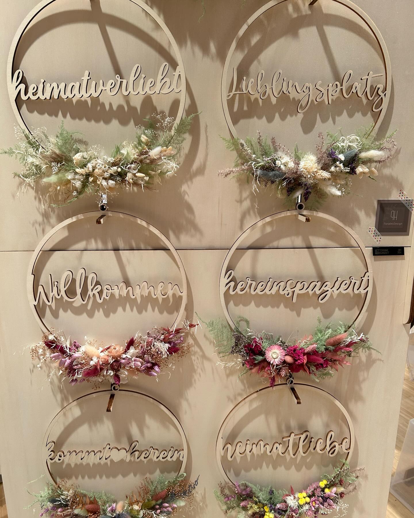Die Abbildung zeigt sechs hölzerne Dekorationen, die an einer Wand hängen. Jede Dekoration besteht aus einem Kranz mit einem Schriftzug und einem Blumengesteck.