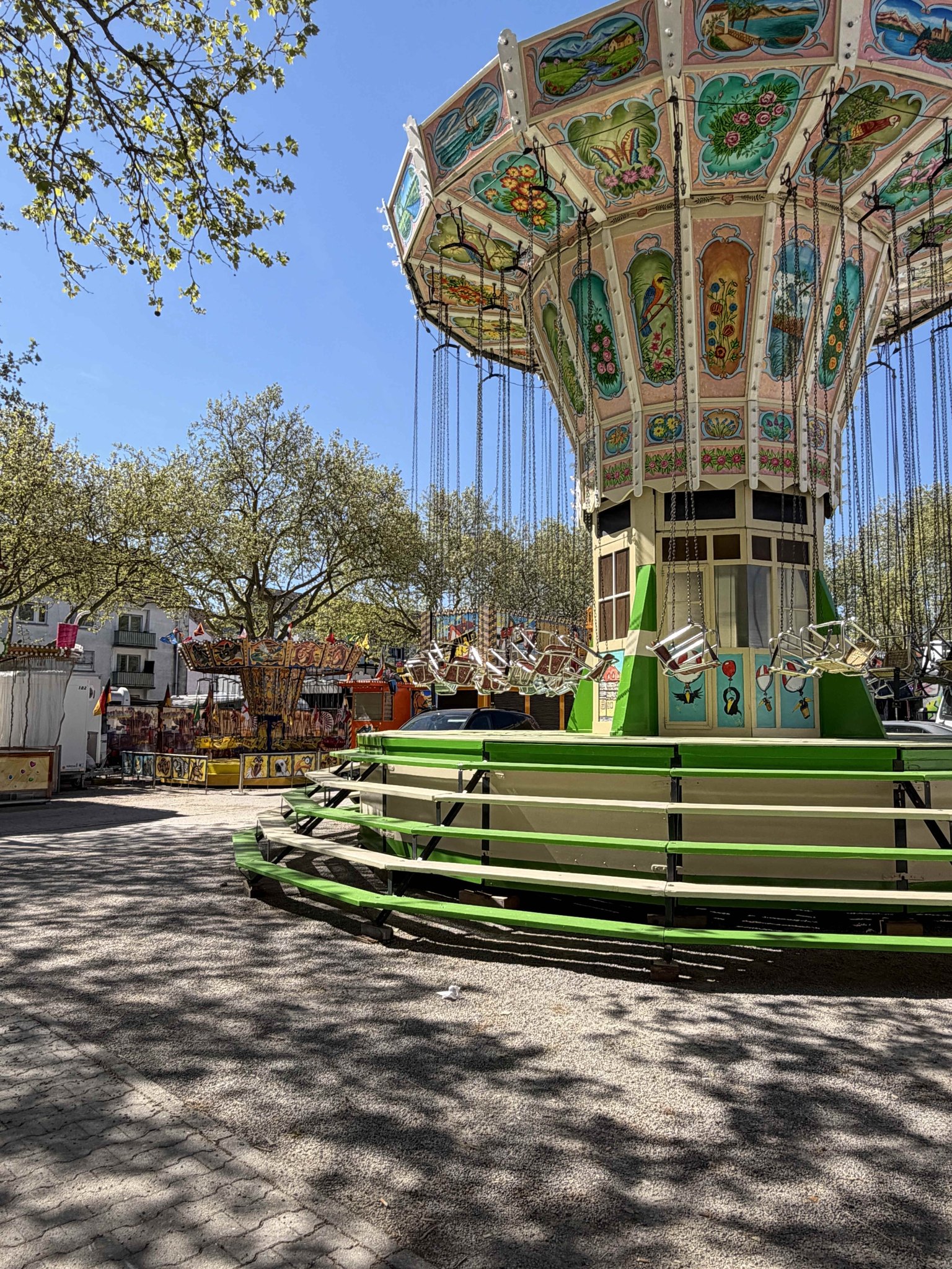 Dieser Alternativtext wurde KI-generiert: Ein bunt verzierter Karussell hängt von einer zentralen Säule herab. Im Hintergrund sind Bäume mit frischem Grün und weitere Fahrgeschäfte zu sehen.