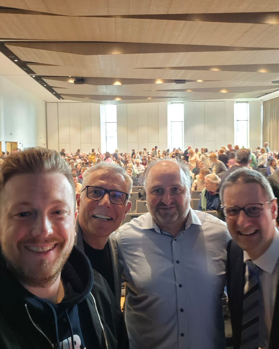 Dieser Alternativtext wurde KI-generiert: Eine Aufnahme von mehreren Personen im Nahbereich vor einer großen Menschenmenge, die in einem hellen Raum mit vielen Fenstern sitzen. Der Hintergrund zeigt eine Bühne und viele Reihen von Stühlen.