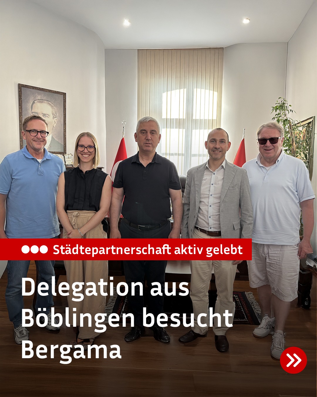 Fünf Personen stehen nebeneinander in einem Raum, im Hintergrund sind Flaggen zu sehen. Ein roter Balken trägt die Aufschrift "Städtepartnerschaft aktiv gelebt". Darunter steht in weißer Schrift "Delegation aus Böblingen besucht Bergama". (KI-Alternativtext)