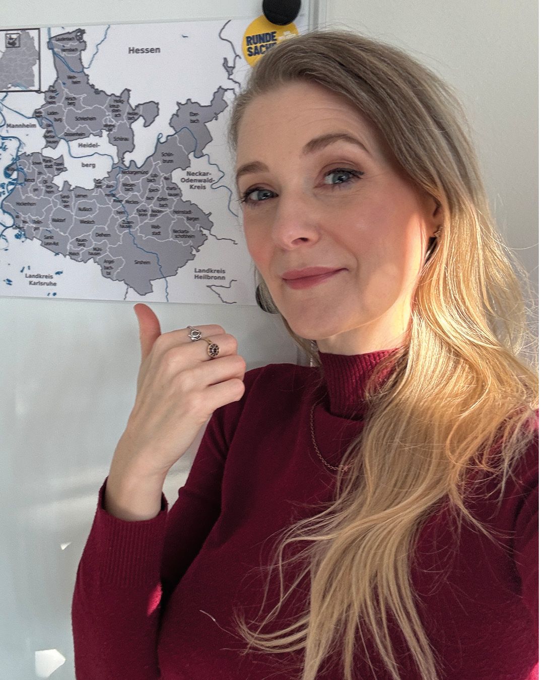 Dieser Alternativtext wurde KI-generiert: Eine Person steht vor einer Karte von Hessen und zeigt einen Ring. Das Bild ist farbig und zeigt eine Innenraumaufnahme.