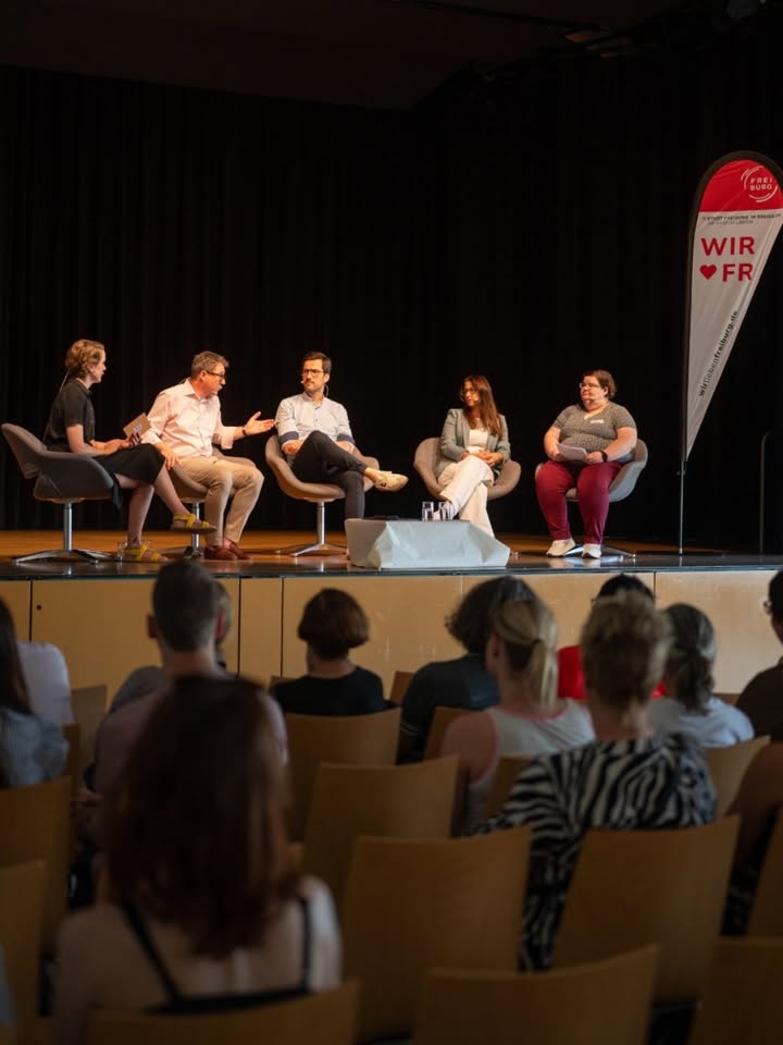 Fünf Personen sitzen auf Stühlen auf einer Bühne vor einem Publikum, eine davon gestikuliert beim Sprechen. Rechts im Bild steht ein Werbebanner mit der Aufschrift „WIR ♥ FR“. Im Vordergrund sind die Köpfe von Zuschauern in Reihen zu sehen. (KI-Alternativtext)