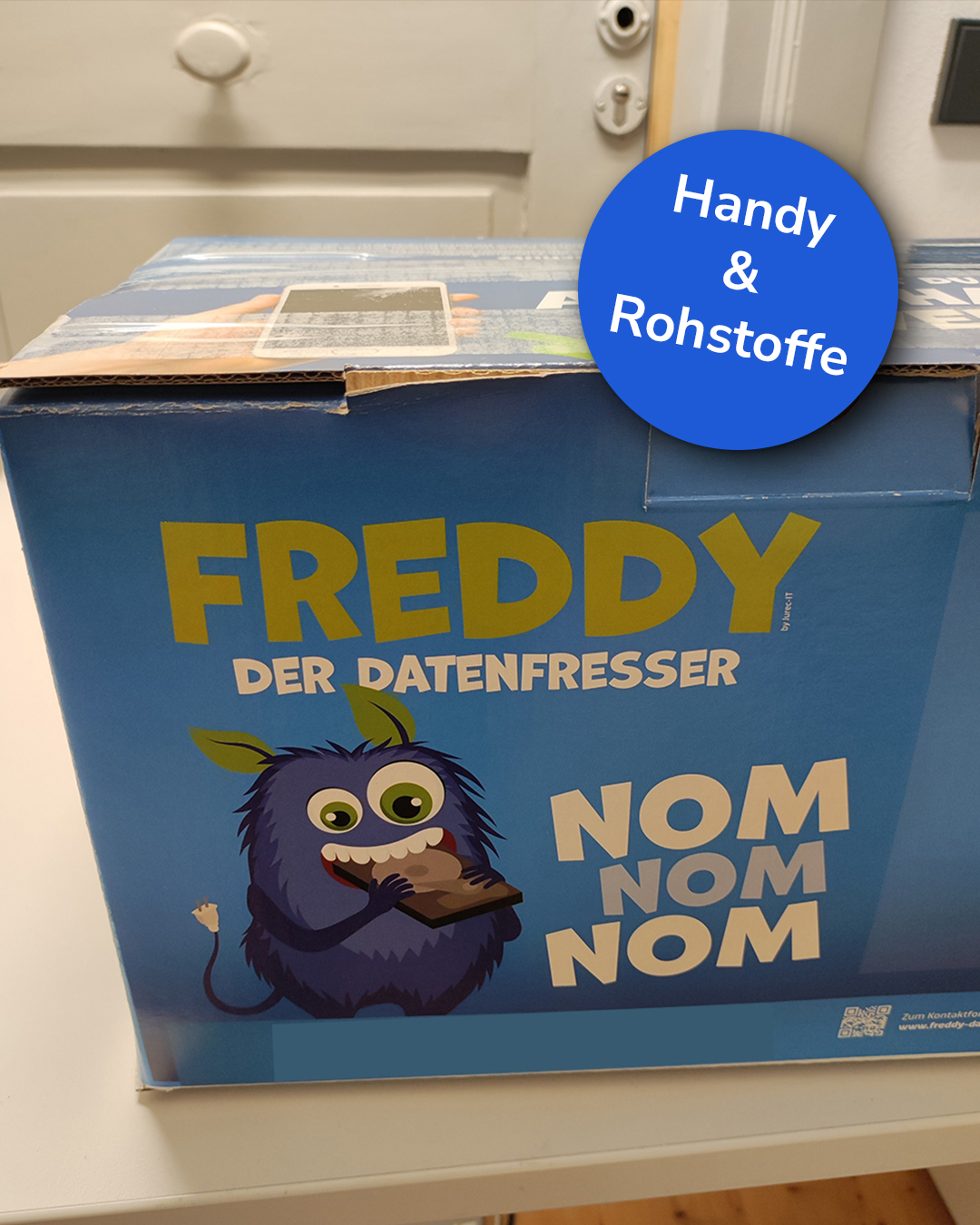 Dieser Alternativtext wurde KI-generiert: Ein blauer Karton steht auf einem Holztisch. Auf dem Karton befinden sich farbenfrohe Illustrationen und Schriftzüge.