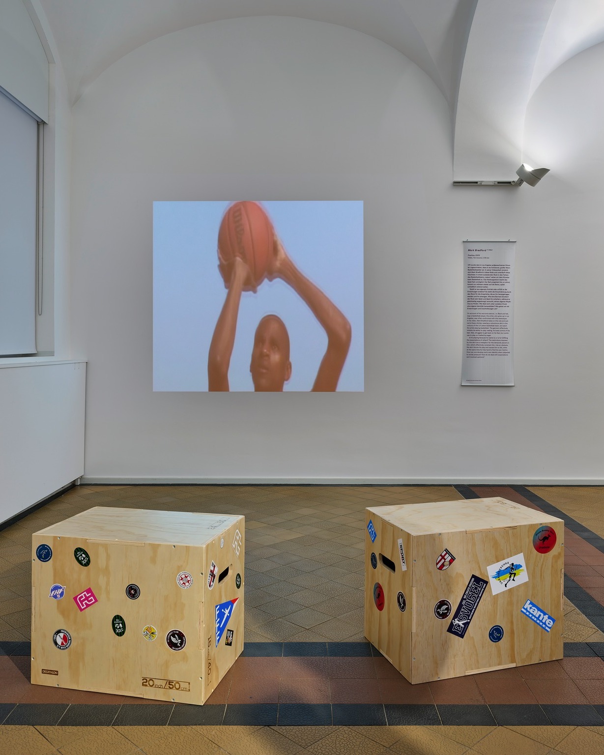 Zwei hölzerne Boxen, bedeckt mit verschiedenen Aufklebern, stehen auf dem gefliesten Boden eines Ausstellungsraums. An der weißen Wand dahinter ist eine Projektion eines Menschen zu sehen, der einen Basketball über dem Kopf hält. Rechts neben der Projektion befindet sich ein weißes Textschild mit dem Titel „Mark Bradford“. (KI-Alternativtext)