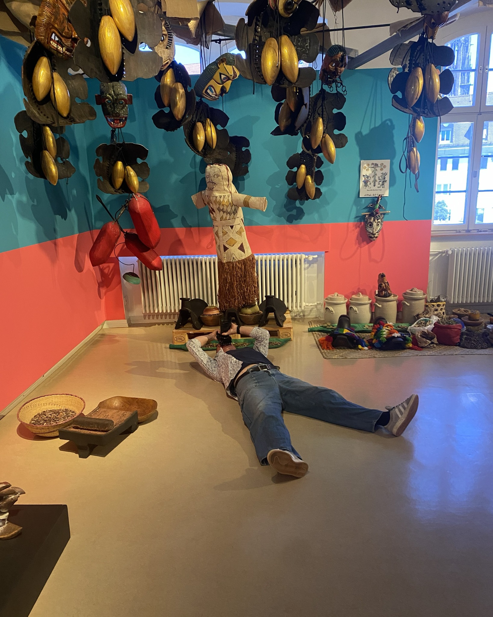 Eine Person liegt auf dem Boden und fotografiert eine stehende Skulptur mit Maskengesicht und Grasrock. Der Raum ist mit zahlreichen hängenden Masken und goldenen, schotenartigen Objekten dekoriert, während auf dem Boden Keramikgefäße und ein Korb mit Steinen stehen. Die Wände sind in Blau und Rot gestrichen. (KI-Alternativtext)