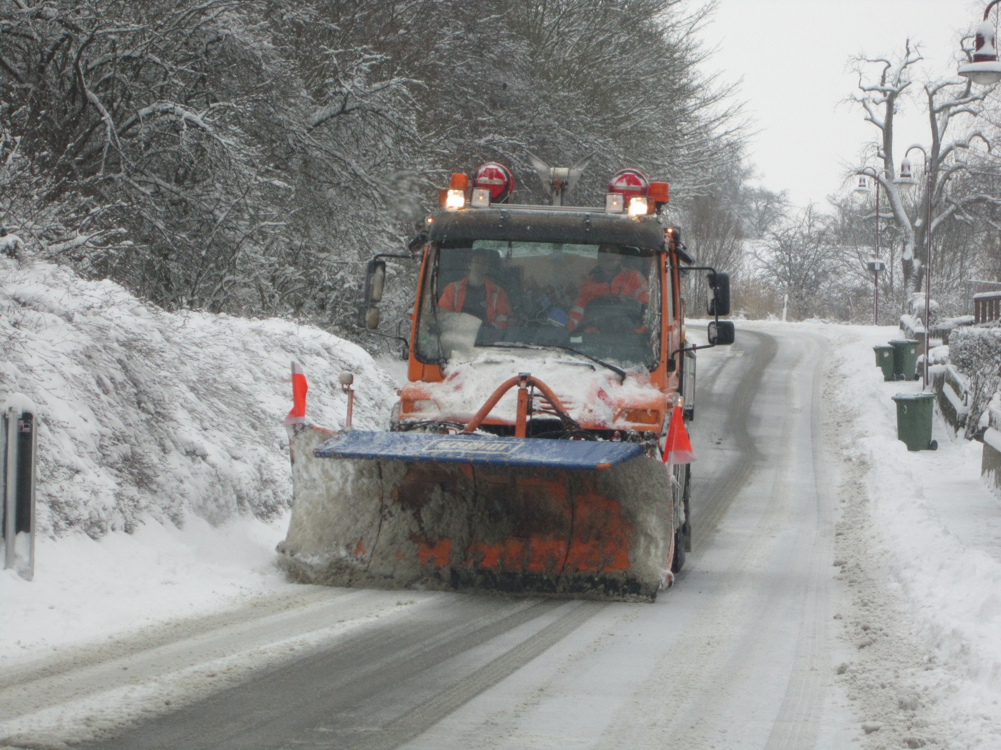 Dieser Alternativtext wurde KI-generiert: Ein orangefarbenes Schneeräumfahrzeug räumt eine verschneite Straße frei. Im Hintergrund sind Bäume und weitere Gegenstände im Schnee zu sehen.