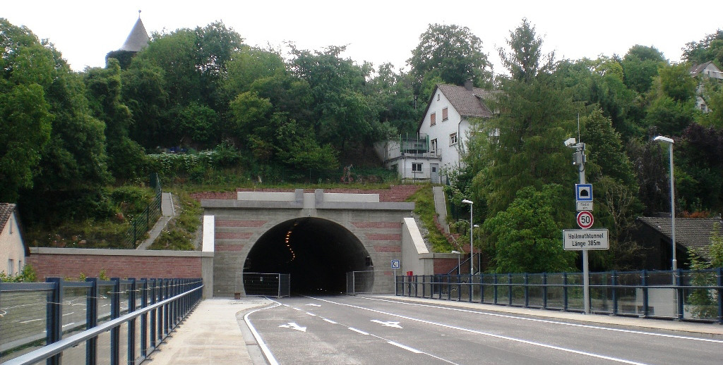 Dieser Alternativtext wurde KI-generiert: Ein Tunnel führt durch einen Hügel, über ihm steht ein Gebäude mit roten Ziegeln. Bäume und Verkehrsschilder befinden sich in der Umgebung.