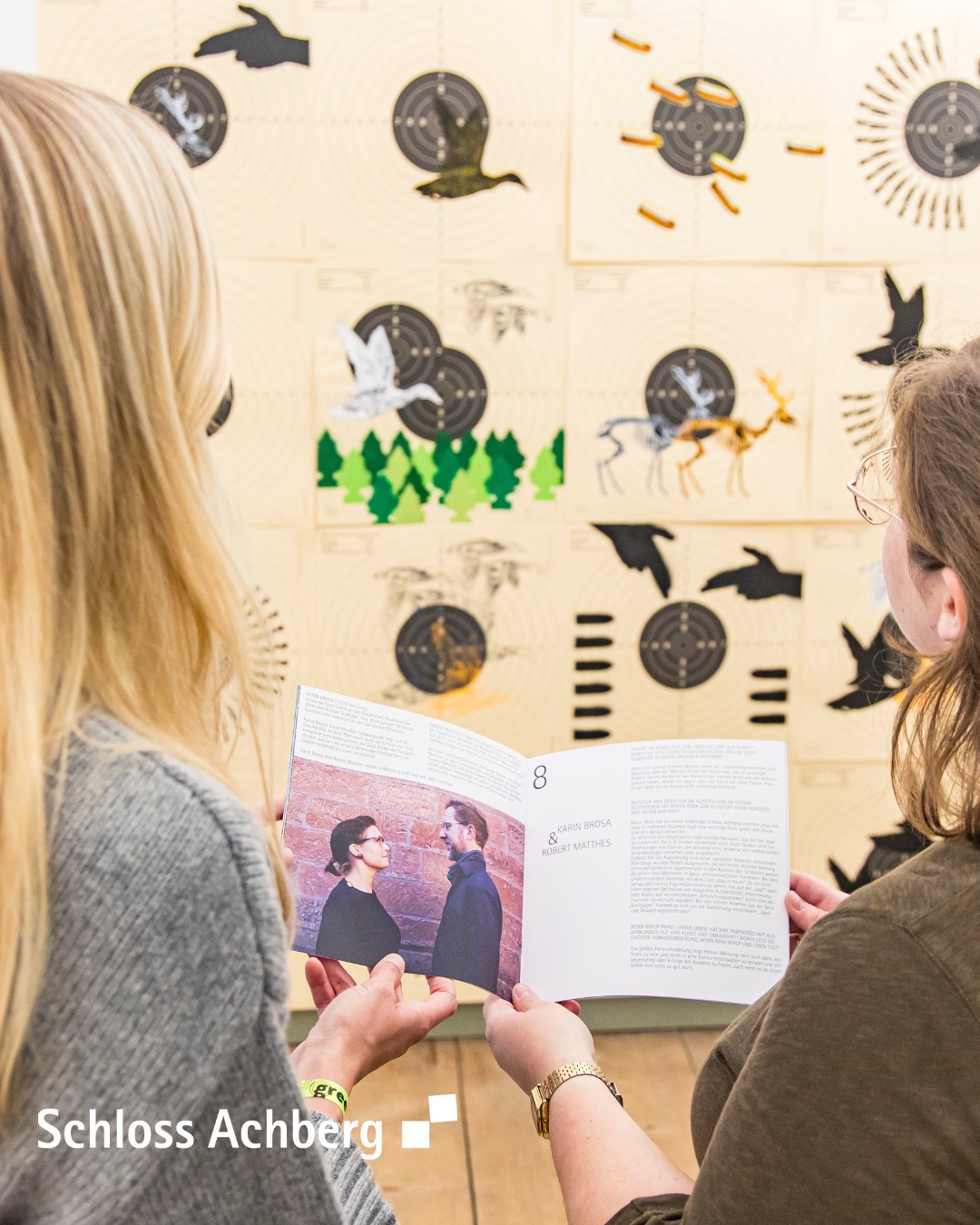 Dieser Alternativtext wurde KI-generiert: Zwei Personen betrachten gemeinsam ein Buch mit Bildern darin. Im Hintergrund befindet sich eine Wanddekoration mit Tier- und Pflanzenmotiven.